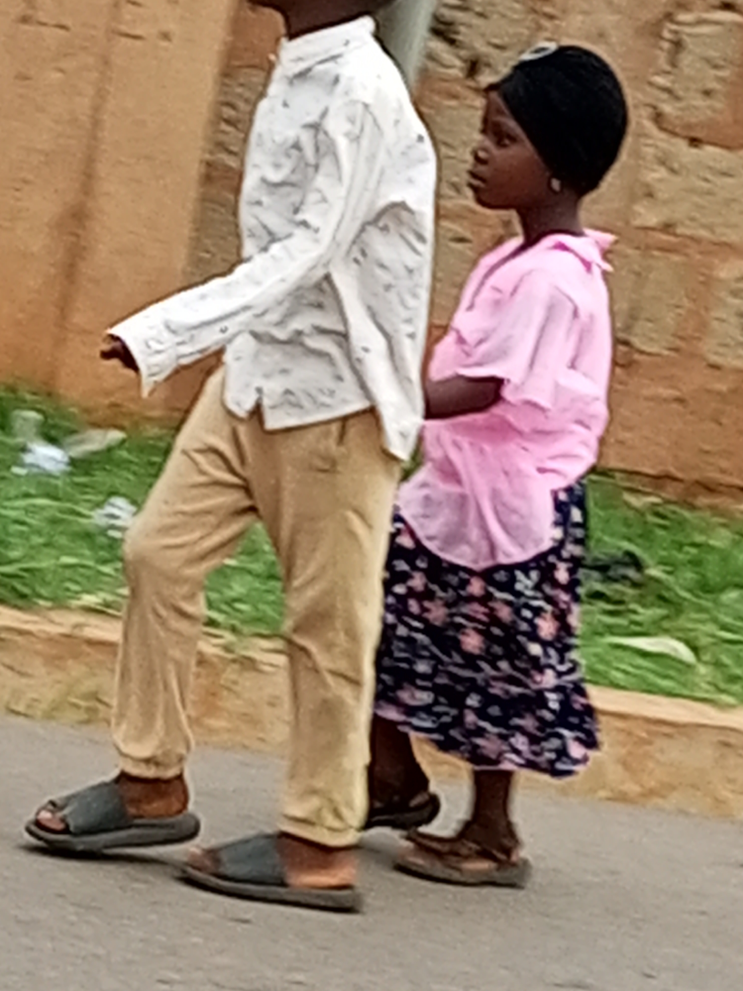 In Tumfure, Nigeria, two individuals, one appearing as an adult male and the other a young female, are captured mid-stride on a paved surface, likely a road. The adult male, positioned on the left, wears a light-colored, patterned long-sleeved shirt tucked into light brown trousers. His footwear consists of dark-colored sandals. The young female, to the right, is dressed in a bright pink, short-sleeved top and a dark, floral-patterned skirt. She also wears dark sandals and a black head covering. Behind them, a strip of green grass runs along a light brown, textured wall, possibly constructed from mud or stone. The lighting suggests it is daytime, and the overall scene depicts a casual street environment.