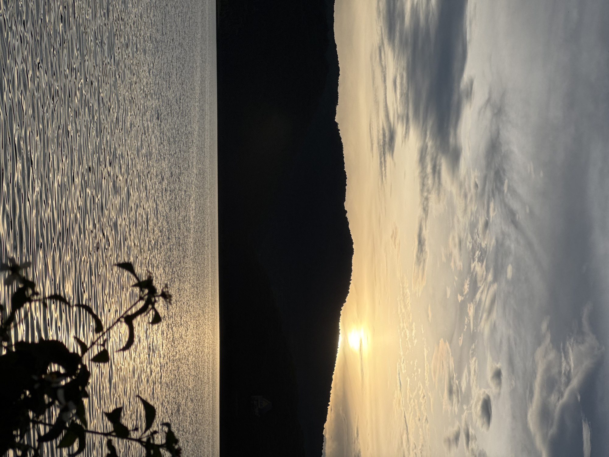 The image depicts a landscape at sunset, likely taken near a body of water in Da Lat, Vietnam.
Environment: The scene is dominated by a body of water, possibly a lake, with small ripples reflecting the sunlight. The right side of the image reveals a silhouette of a mountain range and a cloudy sky with a gradient of colors from yellow-orange near the setting sun to grays and blues higher up.
Foreground: Silhouetted foliage is visible in the bottom left corner.
Overall Impression: The image captures the serene atmosphere of a sunset over a landscape, showcasing the contrast between the bright, reflective water, the dark silhouette of the mountains, and the vibrant sky. The lighting suggests a time around sunset.