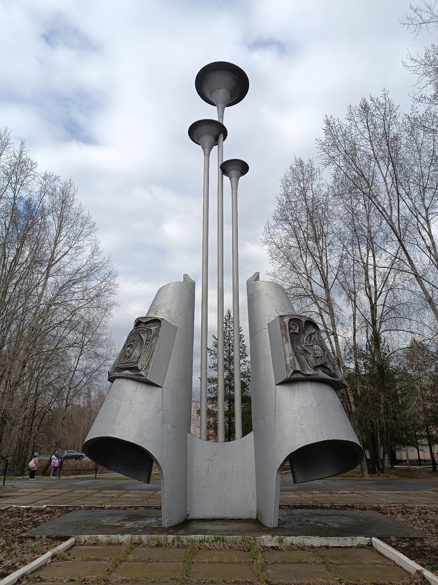 A large, metallic monument stands prominently in what appears to be a public park or square under an overcast sky. The monument features a central cluster of three tall, slender poles, each topped with a disc-shaped element, resembling oversized trumpet bells or stylized light fixtures. These poles rise from between two broad, curved, wing-like structures made of brushed or painted metal, forming the base of the sculpture.
Two rectangular relief sculptures are affixed to the inward-facing surfaces of the curved base sections. The plaque on the left displays a circular emblem, likely the State Emblem of the Soviet Union, accompanied by Cyrillic text. Visible inscriptions include "СОЮЗ ССР" (USSR) above the emblem and "В БОРЬБЕ ЗА МИР" (In the Struggle for Peace) below it. The plaque on the right depicts a figure, possibly a worker or soldier, holding a tool or weapon, with a star and what appears to be a flame. Below this depiction, the Cyrillic word "ОКТЯБРЬ" (October), likely referring to the October Revolution, is visible.
The surrounding environment consists of numerous deciduous trees with bare branches, indicating late autumn, winter, or early spring. A few evergreen trees are also visible in the background. The ground is a mix of concrete paving slabs and patches of earth with sparse grass and fallen leaves. In the distance on the left, two small figures, possibly children, are present, suggesting the area is a public space. A dark-colored car is partially visible in the far background. The time of day is daytime, but the heavy cloud cover creates soft, diffused lighting. The scene is located in Chaikovsky, Russia.