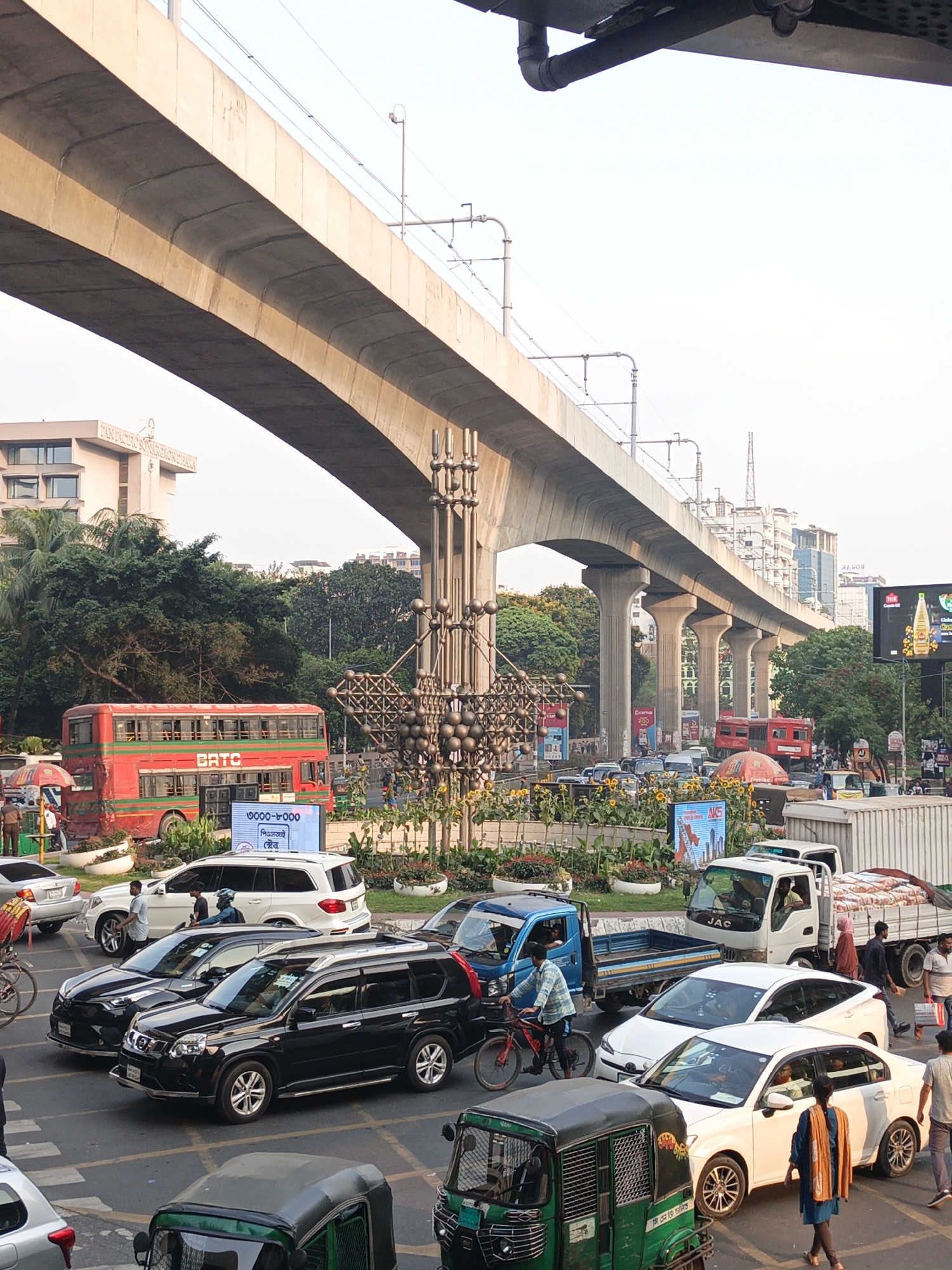 This image captures a bustling street scene in Dhaka, Bangladesh, centered around a multi-lane intersection with heavy traffic and an elevated metro line overhead.
The scene is dominated by the massive concrete structure of an elevated railway or metro line, supported by numerous pillars and featuring overhead power lines. Below it, the street is filled with a dense mix of vehicles, including numerous cars (black and white SUVs, sedans), white trucks (one carrying bags), auto-rickshaws (green, common in Bangladesh), a blue pickup truck, and a red double-decker bus with "BATC" written on its side. A cyclist on a red bicycle is visible among the cars. Pedestrians are seen walking on the sidewalks and attempting to cross the street.
To the left, a multi-story building is partially visible, with "PAN PACIFIC SONARGAON DHAKA" printed on its facade. Lush green trees line parts of the street. In the median of the intersection, there's an elaborate metal sculpture or monument, surrounded by planted flower beds with sunflowers. Several advertising screens or billboards display various texts and images.
The lighting suggests daytime, possibly late afternoon or early evening, with a bright sky overhead and soft light casting shadows. The overall impression is one of a vibrant, active urban environment.
Visible text includes: "PAN PACIFIC SONARGAON DHAKA", "BATC" on the bus, "JAC" on a truck, "AKIS" on a billboard, and Bengali text "৩০০০-৮০০০", "পিএসআই", "স্টোর", "সি এন জি চালিত" (CNG operated), and parts of other advertisements like "Coca-Cola" and "Global".