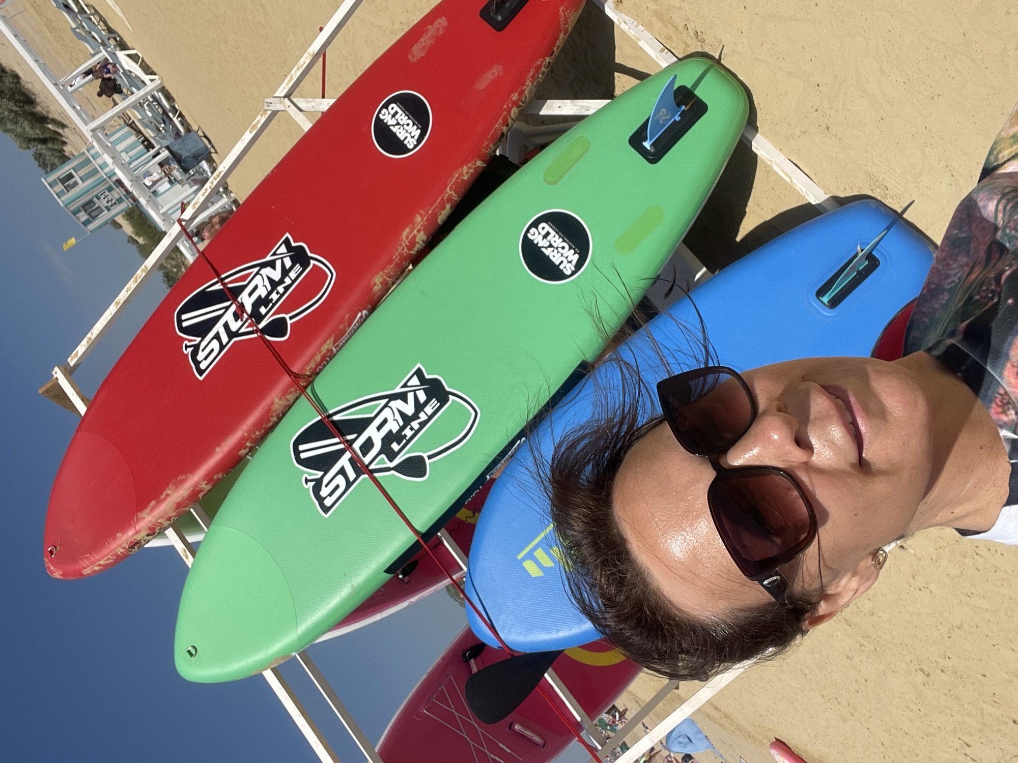 This is a medium shot, eye-level selfie taken on a sandy beach, likely in Anapa, Russia, with a clear blue sky overhead. The main subjects are several stand-up paddleboards (SUPs) arranged on a white rack. From top to bottom, the visible boards are red, green, blue, and a portion of a pink/red board. The red and green boards have black and white logos that read "STORM LINE" and circular stickers with "SURFING WORLD" text. The green board also has a blue fin attached. The blue board has a yellow emblem and a blue fin. In the foreground, a woman is visible from the chest up, wearing large, dark sunglasses, and a patterned shirt. Her hair is dark and windblown. Behind the SUPs, the beach continues with lounge chairs and a small, light-colored building with blue striped accents. The environment appears to be a sunny day at a beach resort.