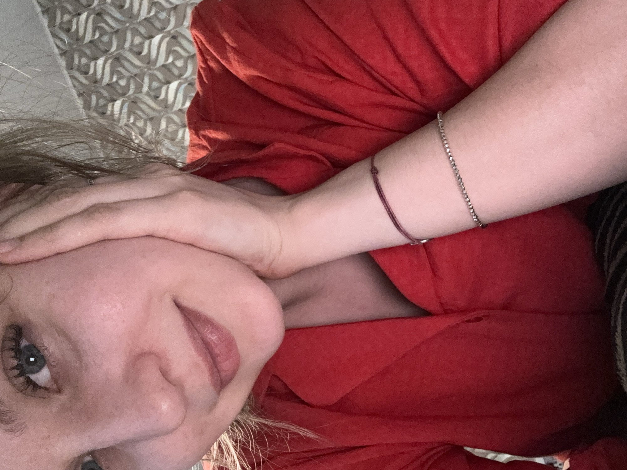 A woman with light skin is lying down, wearing a red shirt. Her left hand is on her forehead, and she is looking at the camera with blue eyes. She has light brown hair. There are two bracelets on her right wrist, one red string and one silver. The background is a patterned surface, possibly a bedspread or pillow. The image does not provide any information about the location in the city of Izhevsk, Russia.