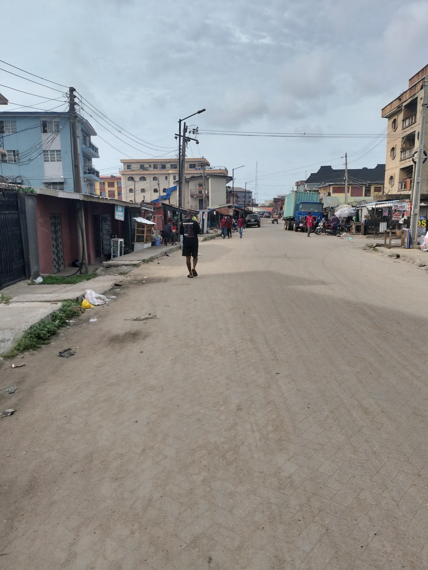The image shows a street in Alaiyabiagba, Nigeria. The road is unpaved, and there is scattered litter. A man walks down the street wearing a black t-shirt with "Pink Panther" printed on the back, and dark shorts. There are buildings on either side of the street, including multi-story structures and single-story shops or stalls. Power lines stretch across the street. In the distance, there are several people and a blue truck. The sky is overcast.