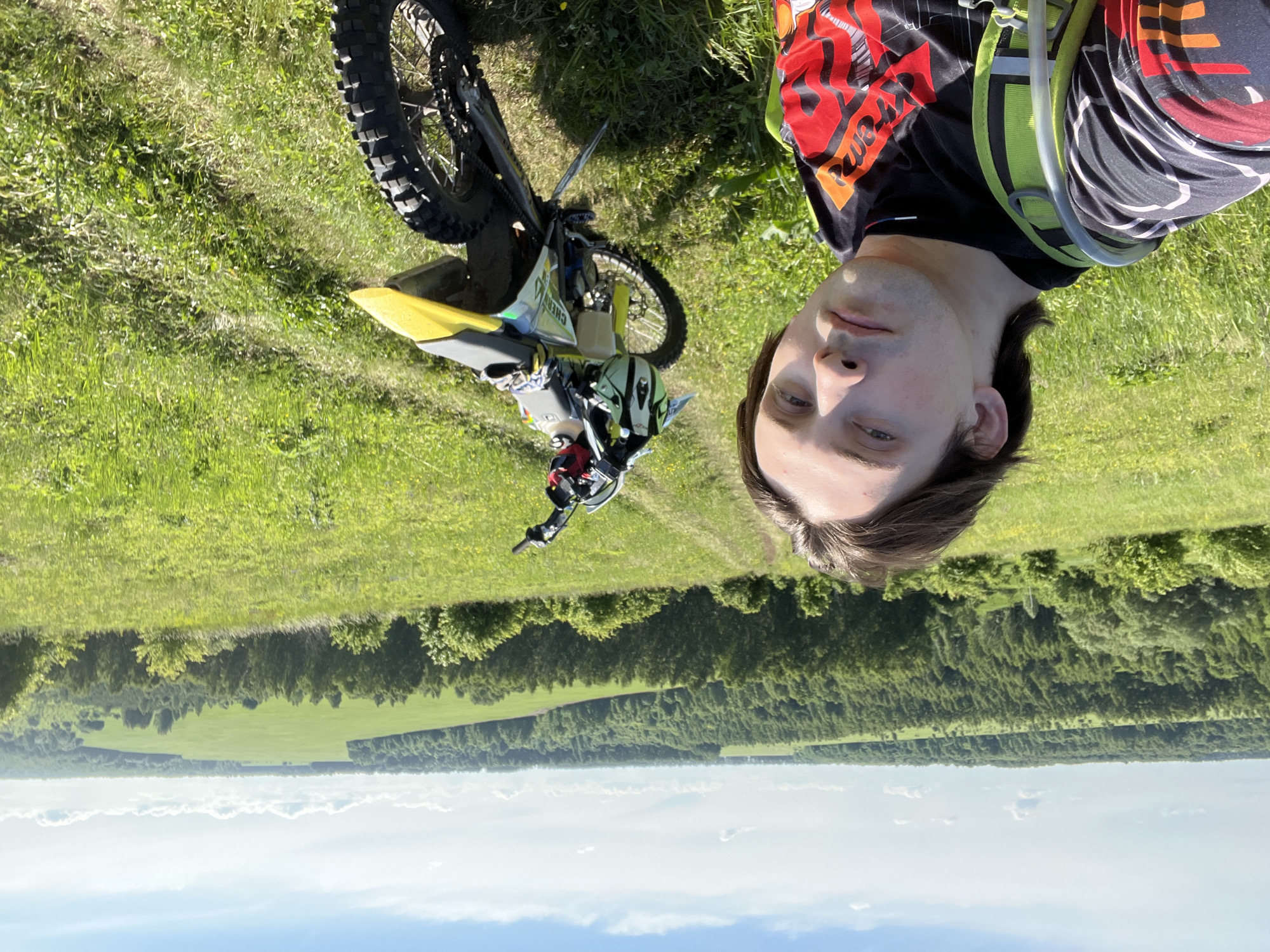 A male individual is captured in a selfie on a sunny day in Vorob'evka, Russia. He stands on a grassy hillside, looking directly into the camera. He has dark, medium-length hair and a serious expression. He wears a dark, patterned long-sleeved shirt with red, orange, and white graphics, and a neon green and black hydration pack or backpack. To his right, a yellow and black dirt bike is parked on a kickstand on a narrow dirt track. A white and green helmet with black goggles, and a pair of gloves, rest on the bike's seat. The track continues into the background, where a vast panorama unfolds. Lush green forests stretch across the midground, beyond which large, bright green cultivated fields are visible. The sky above is clear blue with scattered white cumulus clouds.