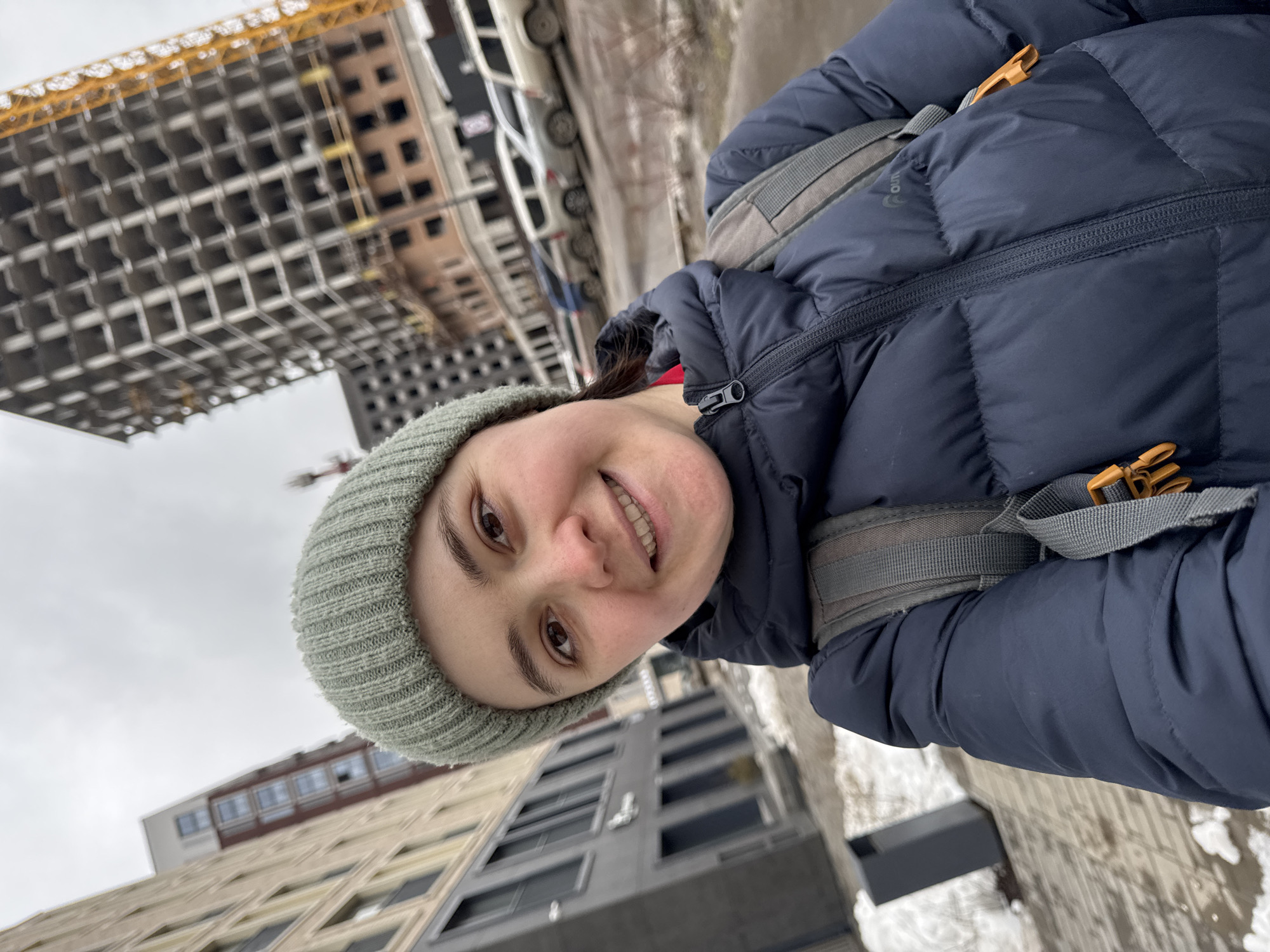 A woman smiles at the camera, wearing a gray-green beanie and a navy blue puffer jacket with a gray backpack. The background features a cityscape with a building under construction, complete with a yellow crane, and other buildings partially visible. The location is Izhevsk, Russia.
