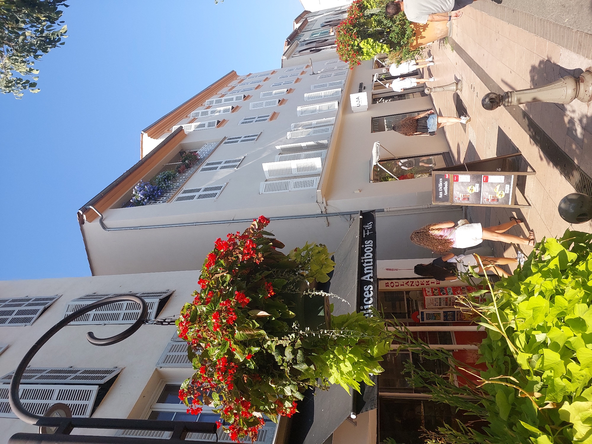 The image shows a sunlit street scene in Antibes, France, likely a commercial area, given the shops visible. In the foreground, lush green foliage is prominent, with a hanging basket filled with vibrant red flowers. Adjacent to this is a storefront with an awning and the text "Aux Délices Antibois Fils" visible on a sign above the entrance. Further down the street, several people are walking on the sidewalk. Two young women in casual summer attire are walking away from the camera. Behind them, two men in white shirts and shorts are walking in the same direction, and further still, a man in a grey t-shirt and shorts is walking towards the camera. The buildings lining the street have a classic Mediterranean architectural style, with light-colored facades, white shutters, and some balconies with flowers. The sky is clear and blue. The street surface appears to be paved with cobblestones or similar material, and there are decorative bollards along the sidewalk.