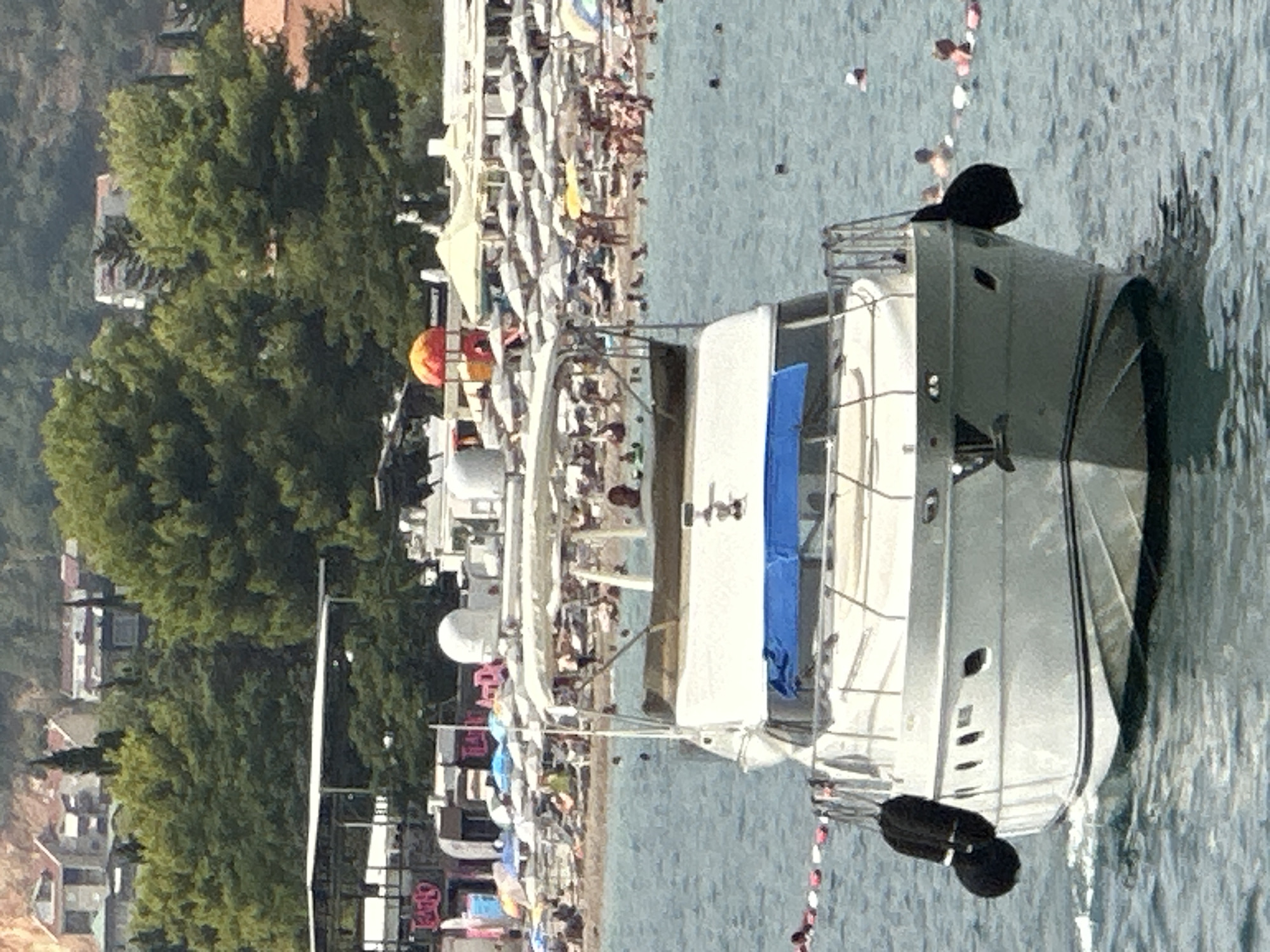 A white motor yacht is docked in the foreground, facing the viewer. The yacht has two black fenders on its port side and a blue tarp covering part of the deck. In the background, a crowded beach is visible with many people sunbathing and swimming in the sea. Beach umbrellas and some buildings are also present on the shore. The scene appears to be a sunny day at a popular tourist destination. Based on the surrounding architecture and the overall ambiance, the location is likely Budva, Montenegro.