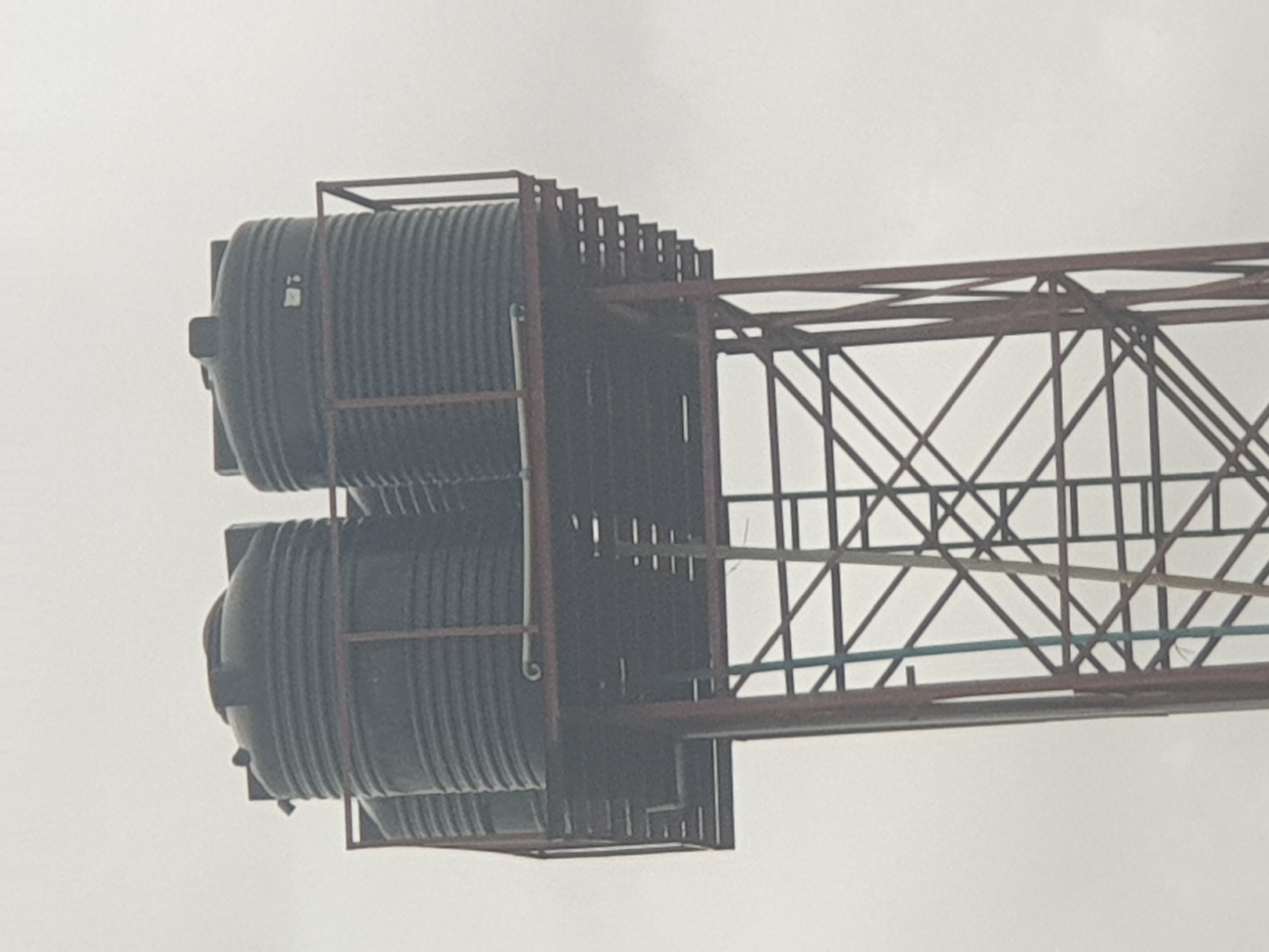 An outdoor, waist-level, slightly blurry, medium shot captures a tall, rusty metal water tower supporting three large, black plastic water tanks against a pale, overcast sky. The tower has a crisscross framework of metal beams. The water tanks are cylindrical with ridged sides and dark lids. One tank is partially obscured by another. There are pipes running from the tanks to the ground, and a narrow metal walkway or ladder is attached to the side of the tower. The scene is devoid of people, animals, or any vegetation. The image appears to be taken in Jalingo, Nigeria.