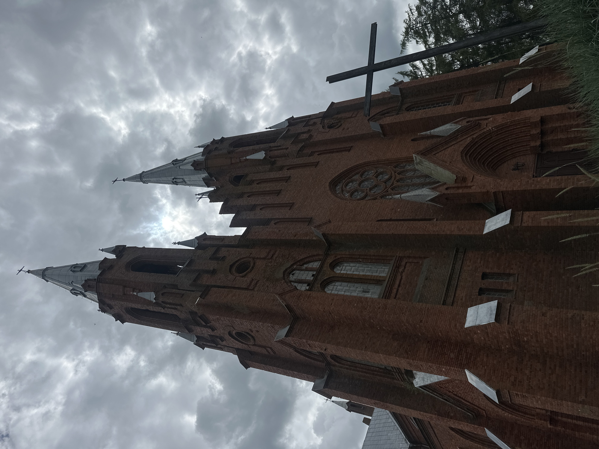 The photograph captures a gothic-style brick church with twin spires reaching towards a cloudy, overcast sky. The church is constructed from dark red bricks and features tall, arched windows, a rose window, and multiple pointed architectural elements. In the foreground, a large, dark wooden cross stands to the right of the church, silhouetted against the building and some distant trees. The ground in the immediate foreground is covered with tall, dry grass. The overall atmosphere is somber and dramatic due to the dark clouds and low-angle perspective of the building.