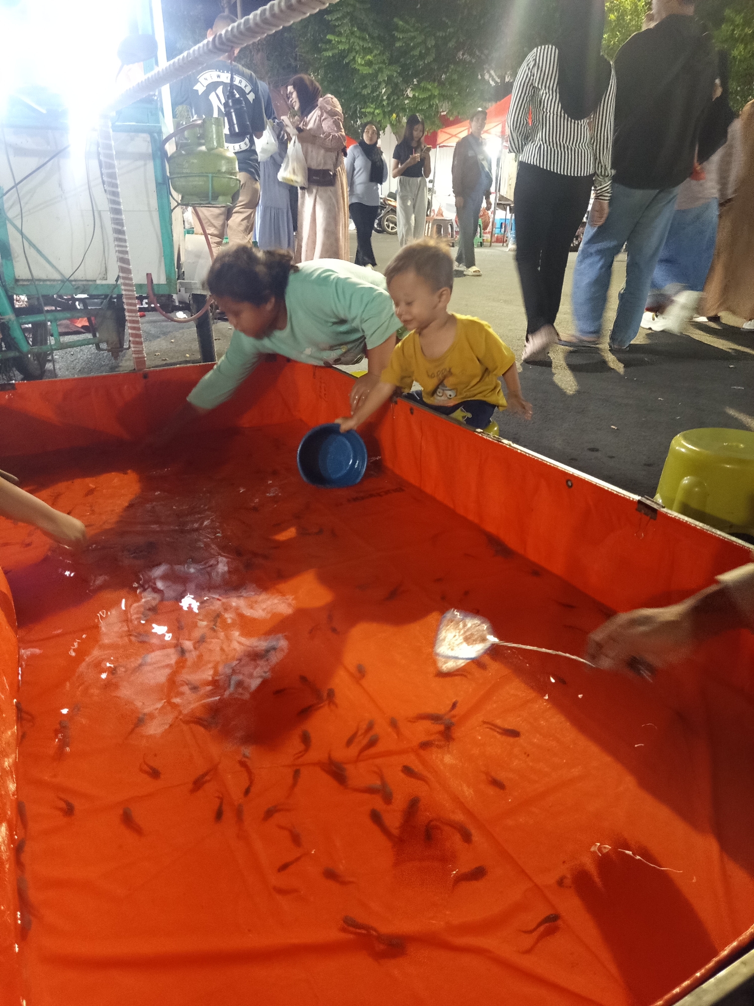 An outdoor scene depicts a game of goldfish catching at night. A large, bright orange tub filled with water and small fish is the central focus. Two children are actively participating in the game. A young boy wearing a yellow shirt and blue shorts is reaching into the tub, and a young girl in a green shirt is also attempting to catch fish. Other people, including adults, are visible in the background, some observing the game. The environment appears to be a street or public space, with a food cart or stall visible on the left, illuminated by bright lights, suggesting a festive atmosphere. The presence of a green gas cylinder on the cart points to a food preparation area. The ground is paved, and trees are visible in the background, indicating an outdoor urban setting. The overall lighting suggests it is nighttime, possibly during a local fair or event in Situbondo, Indonesia, a coastal city known for its festivals.
