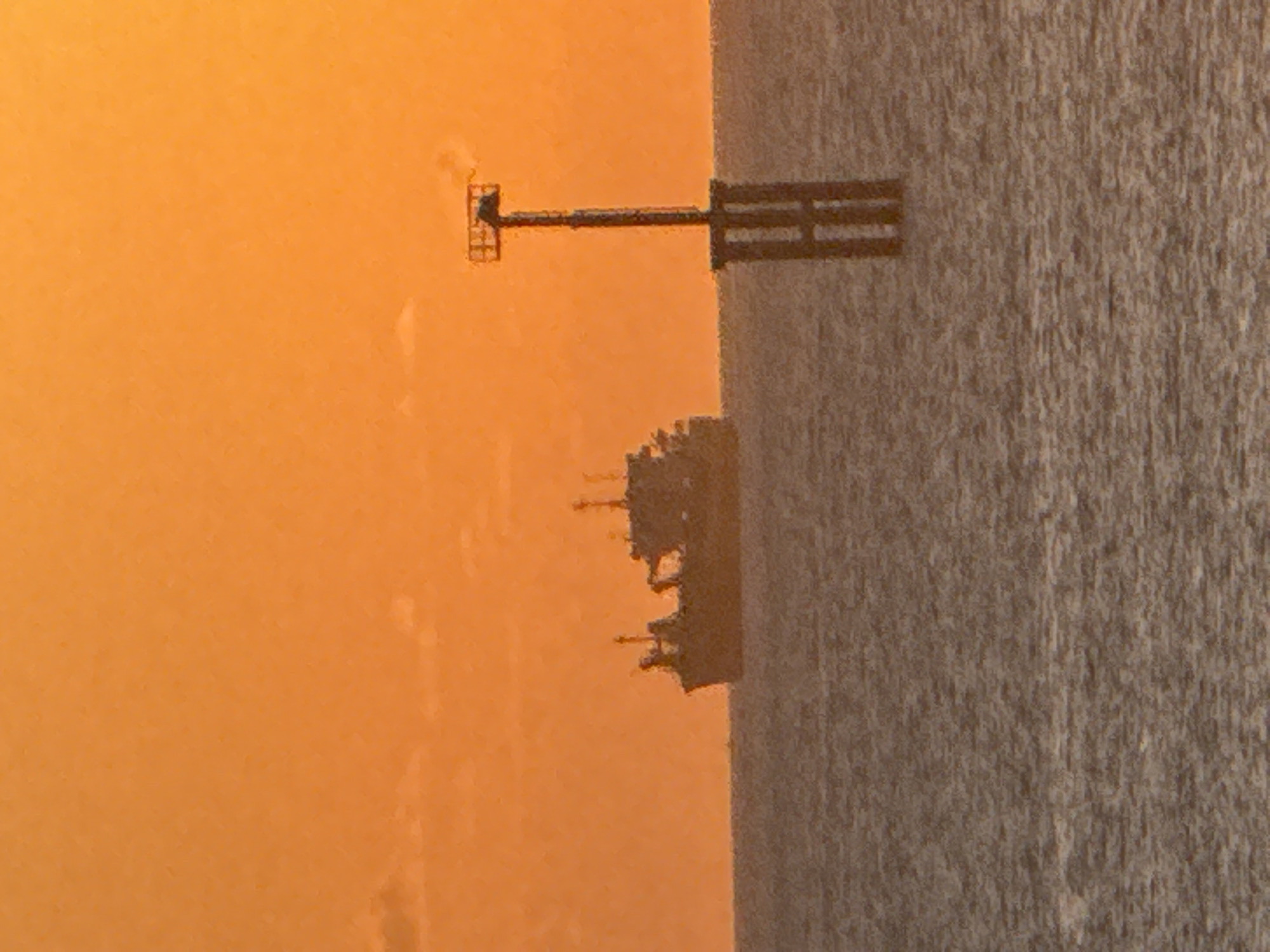 The image depicts a serene coastal or marine scene during either sunrise or sunset, casting a vibrant orange glow across the sky. In the foreground, a dark, textured vertical surface occupies the right side of the frame, resembling a wooden or concrete structure with a coarse, grainy pattern.
Extending horizontally from this right-hand structure is a dark, silhouetted pier or jetty. It stretches into the bright orange backdrop, culminating in a small, elevated platform enclosed by railings. Below this pier, closer to the right side, a more complex and irregular silhouette is visible. This distant outline suggests an industrial facility or an offshore structure like an oil rig, possibly part of a distant coastline or port.
The warm, atmospheric lighting creates a tranquil and somewhat mysterious mood. There are no visible people or discernible activities taking place. No text is visible within the image. Given the context of Kuta, Indonesia, the scene likely captures a coastal view with man-made structures against a dramatic sky.