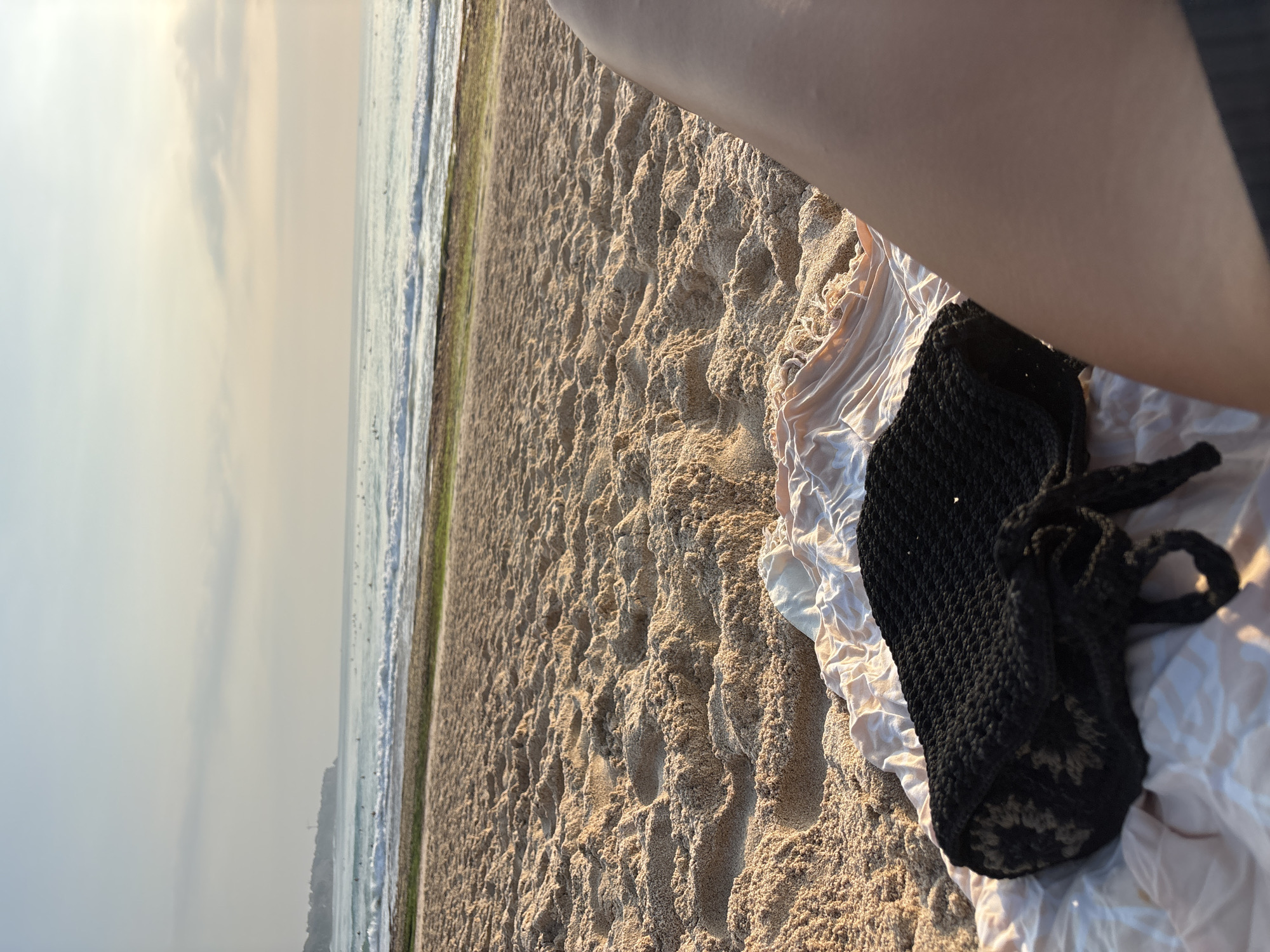 The image shows a beach scene in Kuta Selatan, Indonesia. The foreground features a black crocheted bag placed on a cream-colored throw. To the right of the bag, a person's leg is visible, suggesting they are sitting or lying on the beach. The sand is textured with footprints and ripples. In the middle ground, the ocean stretches out, with waves gently breaking near the shore. Several surfers are scattered across the water, indicating activity. The background shows a hazy coastline with a faint cliff formation and a lighthouse or tower. The sky is overcast with soft clouds, and the overall lighting suggests either early morning or late afternoon.