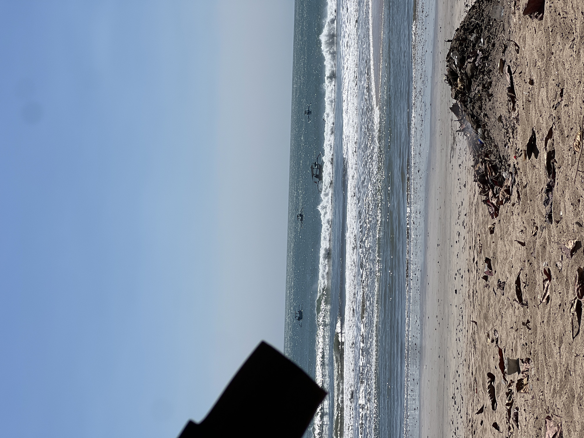 The image depicts a beach scene in Kuta, Indonesia, with the ocean and sky dominating the background. In the foreground, a pile of debris, likely trash, is visible on the sandy beach, with some smoke emanating from it, suggesting it might be smoldering. Scattered dry leaves and debris are also present on the sand. The ocean shows moderate waves breaking on the shore, with several small boats with outriggers visible in the water. The sky is clear blue, indicating a sunny day. A dark, angular object, possibly part of a structure or object not fully in frame, occupies the top left portion of the image.