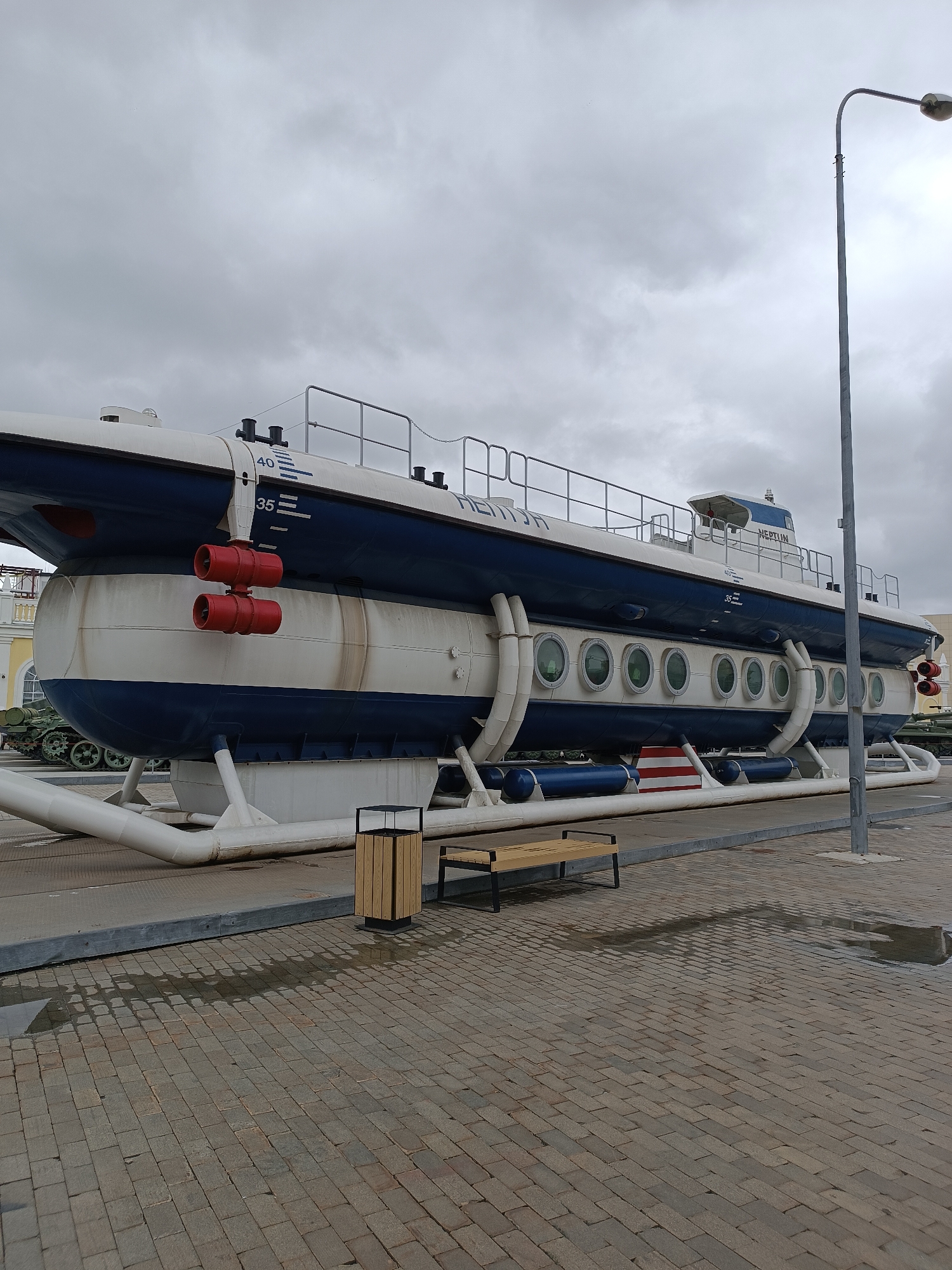 In the city of Verkhnyaya Pyshma, Russia, a museum exhibit features a deactivated submarine displayed outdoors. The submarine, painted in white and dark blue, is elevated on a white support structure. Prominent red cylindrical objects are mounted on its exterior, and a series of round portholes are visible along its side. The submarine bears markings, including the word "NEPTUN" and numerical depth indicators. In the foreground, a paved area with cobblestones, wet from recent rain, features a modern trash receptacle and a wooden bench. Behind the submarine, remnants of military vehicles, possibly tanks, can be seen. The sky above is overcast.
