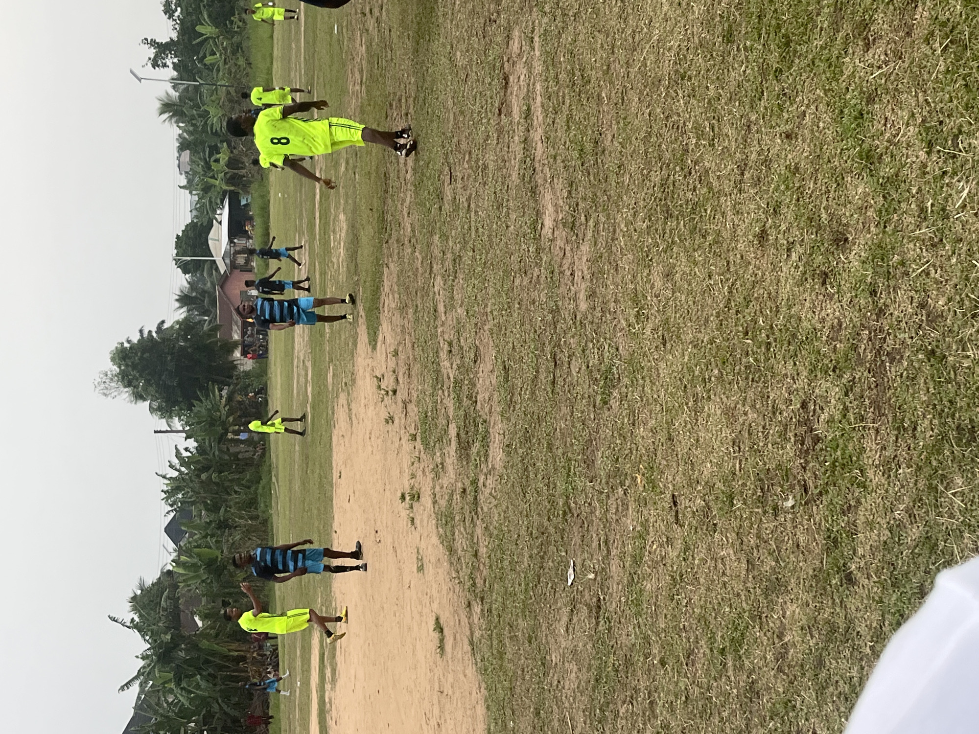 The image shows an outdoor soccer field in Ikot Ekpene, Nigeria. The field is composed of both grass and bare sandy areas. Several players, wearing bright yellow and blue-striped jerseys, are scattered across the field. One player, wearing jersey number 8, is more prominent in the foreground. Trees and buildings are visible in the background. The sky is overcast.