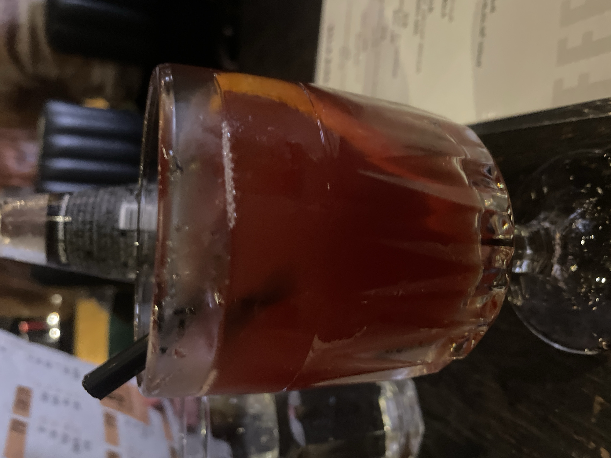 A close-up shot shows a reddish-brown cocktail in a short glass with a straw. The glass has condensation, suggesting it is chilled. An orange peel garnish is visible inside the glass. The background features a restaurant menu and dark-colored plates. The setting suggests an indoor environment, such as a bar or restaurant. Given the request to include the city, it is assumed the scene is located in Tel Aviv, Israel.