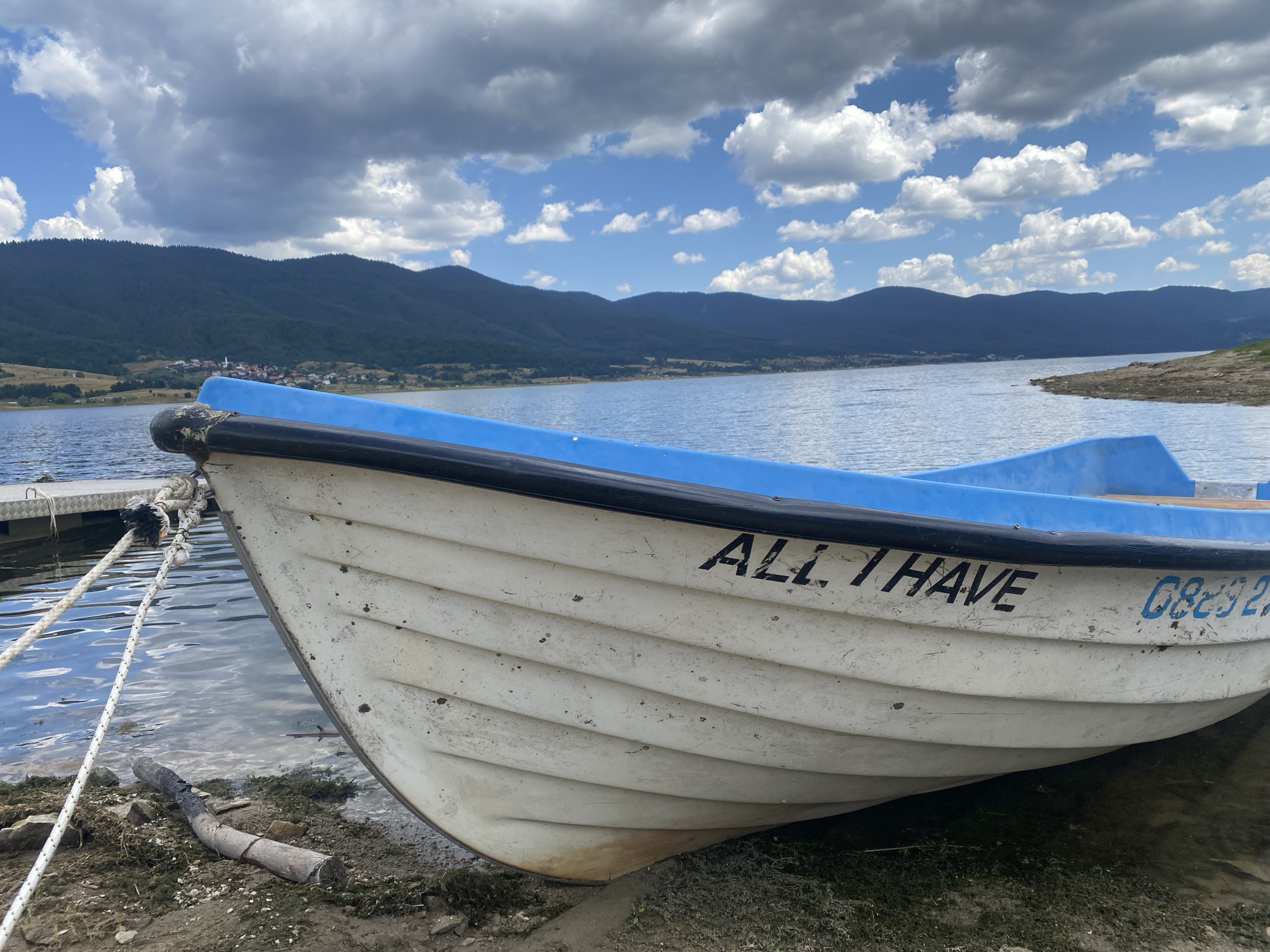 A white rowboat with a blue rim is partially beached on the shore of the Sarnitsa Reservoir in Sarnitsa, Bulgaria. The boat is tied to a dock with a rope. The boat has the name "ALL I HAVE" painted on its side, followed by a registration number that appears to be "GB292". The background shows a body of water, rolling hills covered in trees, and a cloudy sky. The shore is composed of dirt, rocks, and some vegetation. There are no people visible in the image.