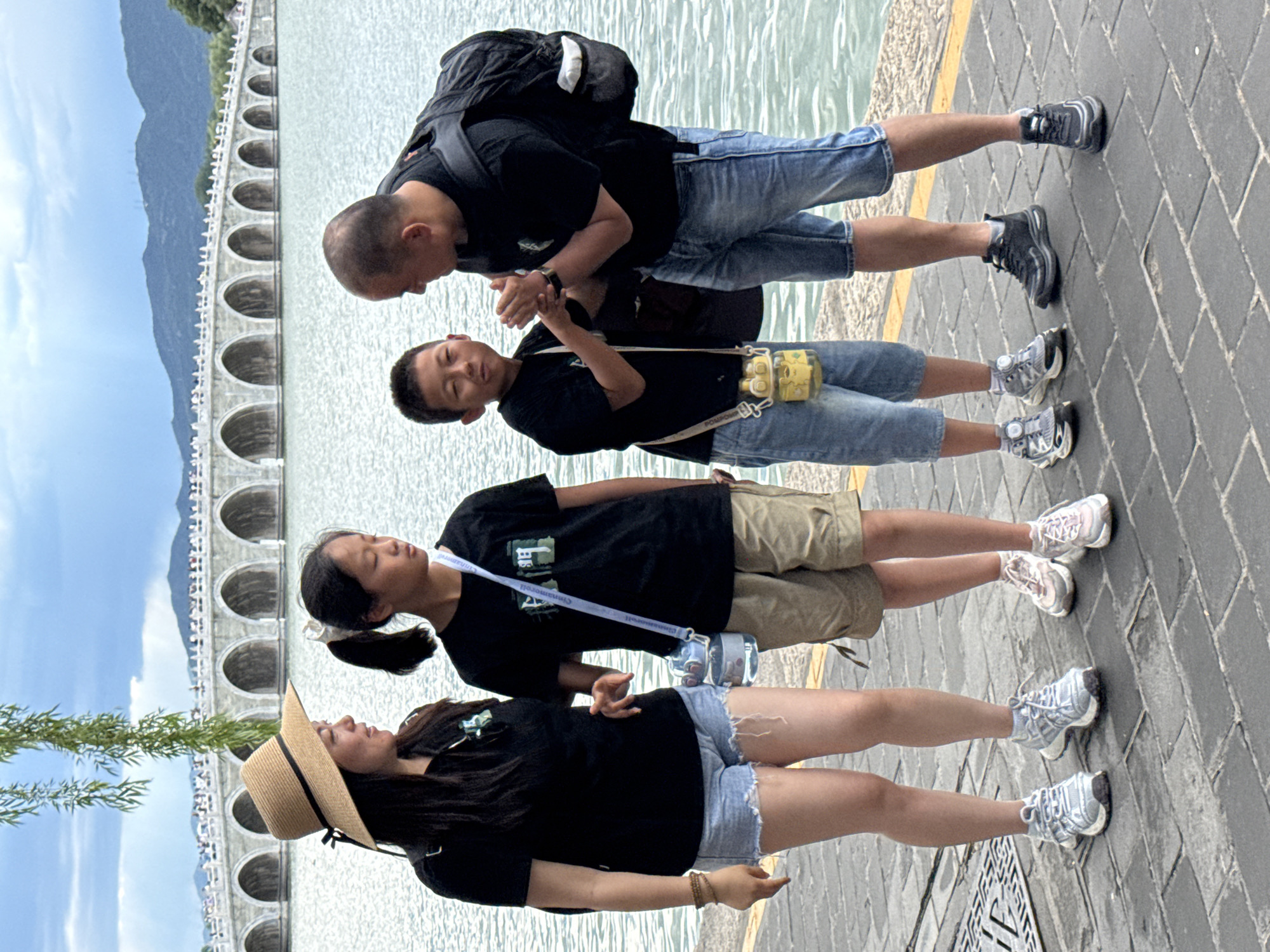 A group of four individuals, consisting of two adults (one female, one male) and two children (one girl, one boy), stands on a grey stone paved walkway adjacent to a large body of water. The background features the 17-Arch Bridge spanning across Kunming Lake in the Summer Palace, Haidian District, Beijing, China, with green mountains and a cloudy sky beyond. A tree branch with green leaves hangs into the frame from the upper left.
The female adult, positioned on the far left, wears a straw sun hat with a black band, a black short-sleeved t-shirt, light blue denim cutoff shorts, and light-colored athletic shoes. A brown beaded bracelet is visible on her left wrist. She looks towards the female child.
The female child, to the right of the female adult, wears a black short-sleeved t-shirt, khaki shorts, and light pink and white athletic shoes. Her dark hair is tied in a ponytail with a white scrunchie. A clear cylindrical water bottle hangs from a white strap around her neck. She looks downwards and slightly to the right.
The male child, to the right of the female child, wears a black short-sleeved t-shirt, light blue denim shorts, and grey and white athletic shoes with yellow accents. A yellow animal-shaped water bottle hangs from a strap around his neck. He is facing the male adult, with his hands clasped together near the male adult's hands.
The male adult, on the far right, has a bald head and wears a black short-sleeved t-shirt, blue denim shorts, and dark athletic shoes. A black backpack is worn on his shoulders. He leans slightly towards the male child, engaging in a hand interaction with the child, his hands also clasped together.
The walkway includes a metal grate on the left and a yellow painted line along its edge on the right. The water surface exhibits minor ripples.