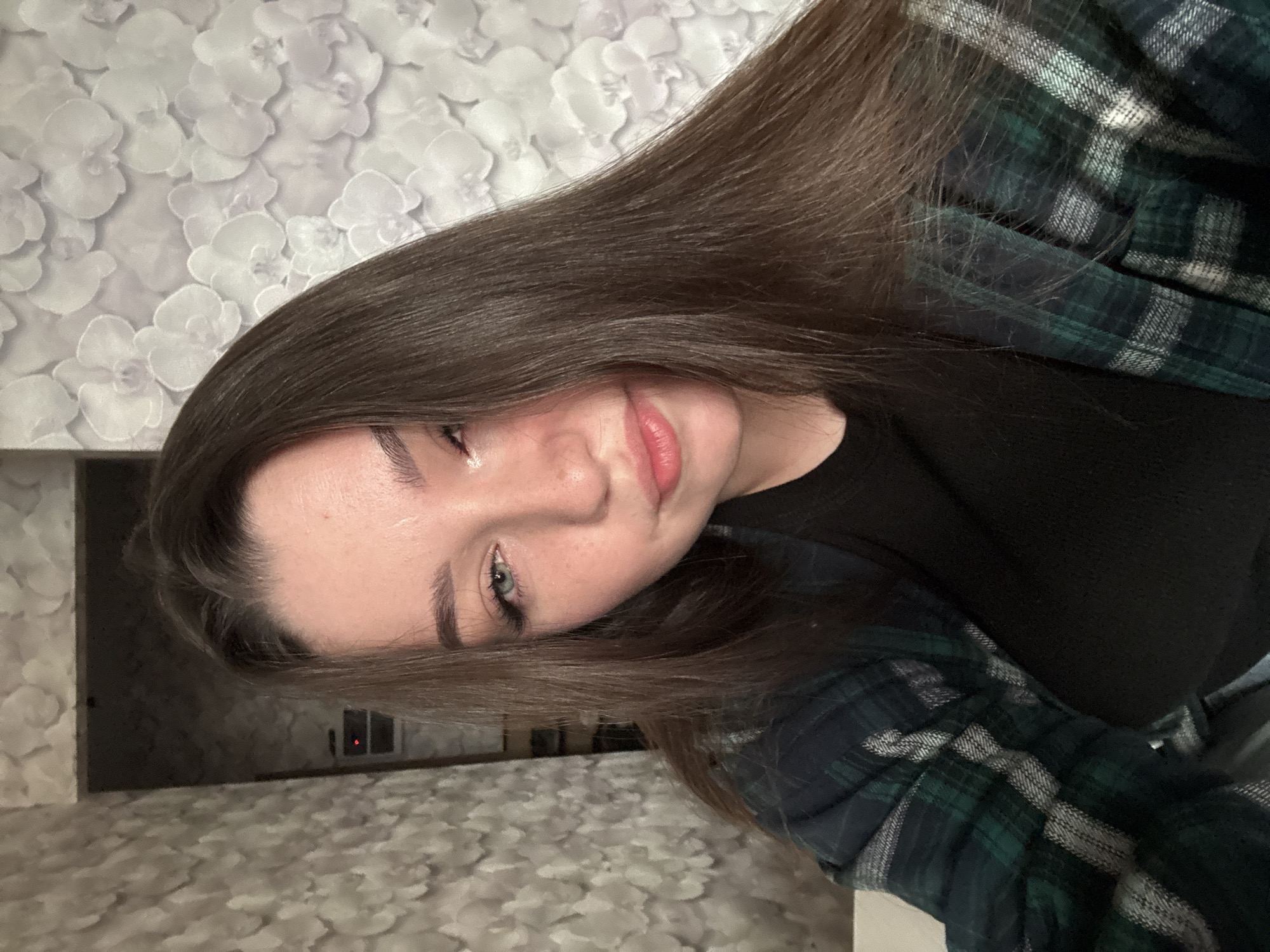 A young woman with long, dark brown hair is shown in a close-up, selfie-style photograph. She is wearing a black t-shirt and an open, dark green and black plaid flannel shirt. Her face is partially turned to the side, with her right eye and lips clearly visible. Her left eye is looking directly at the camera. The background consists of wallpaper with a repeating floral pattern. The image appears to have been taken indoors. The location in the city of Osentsy, Russia, is not identifiable from the visual information provided.