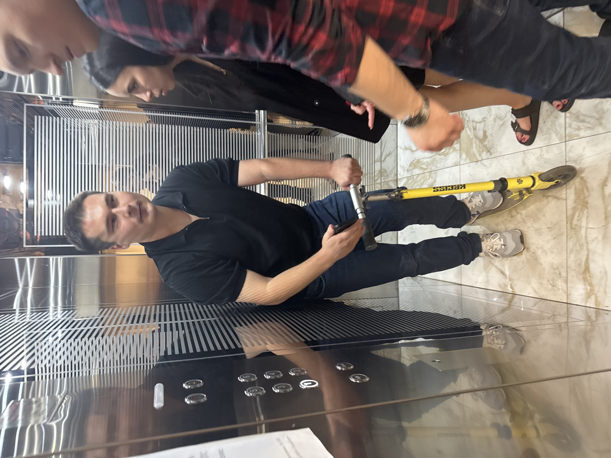 In an elevator, a man in a black polo shirt and jeans stands with his left foot on a yellow scooter, holding its handlebars. To his right, a woman in a black coat and dark pants stands with her left foot in a dark sandal. To the right of the woman, another person, mostly out of frame, wears a red and black plaid shirt and dark pants, with a watch on their left wrist. The elevator walls are made of reflective metal with horizontal striped patterns. The floor is tiled with a marble-like pattern. The man on the scooter is facing the camera, while the woman and the third person are looking towards the man. The scene appears to be an indoor residential or commercial building elevator. The presence of an elevator and specific building finishes do not provide enough information to definitively place the image in the city of Perm, Russia.