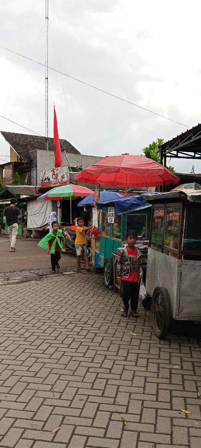 The image depicts a street scene in Banjaran, Indonesia, located at coordinates -7.046 latitude and 107.581 longitude. A man is walking down a brick-paved street. Three children, two boys and a girl, are standing near three food carts. The girl is wearing a green cape and the boys are wearing shirts and shorts. The carts are covered with awnings and have various snacks for sale. One cart is decorated with a sign that reads "Batagor." The street is lined with buildings and a tall tower with a red flag. There are multiple wires running above the street. The sky is overcast and gray.