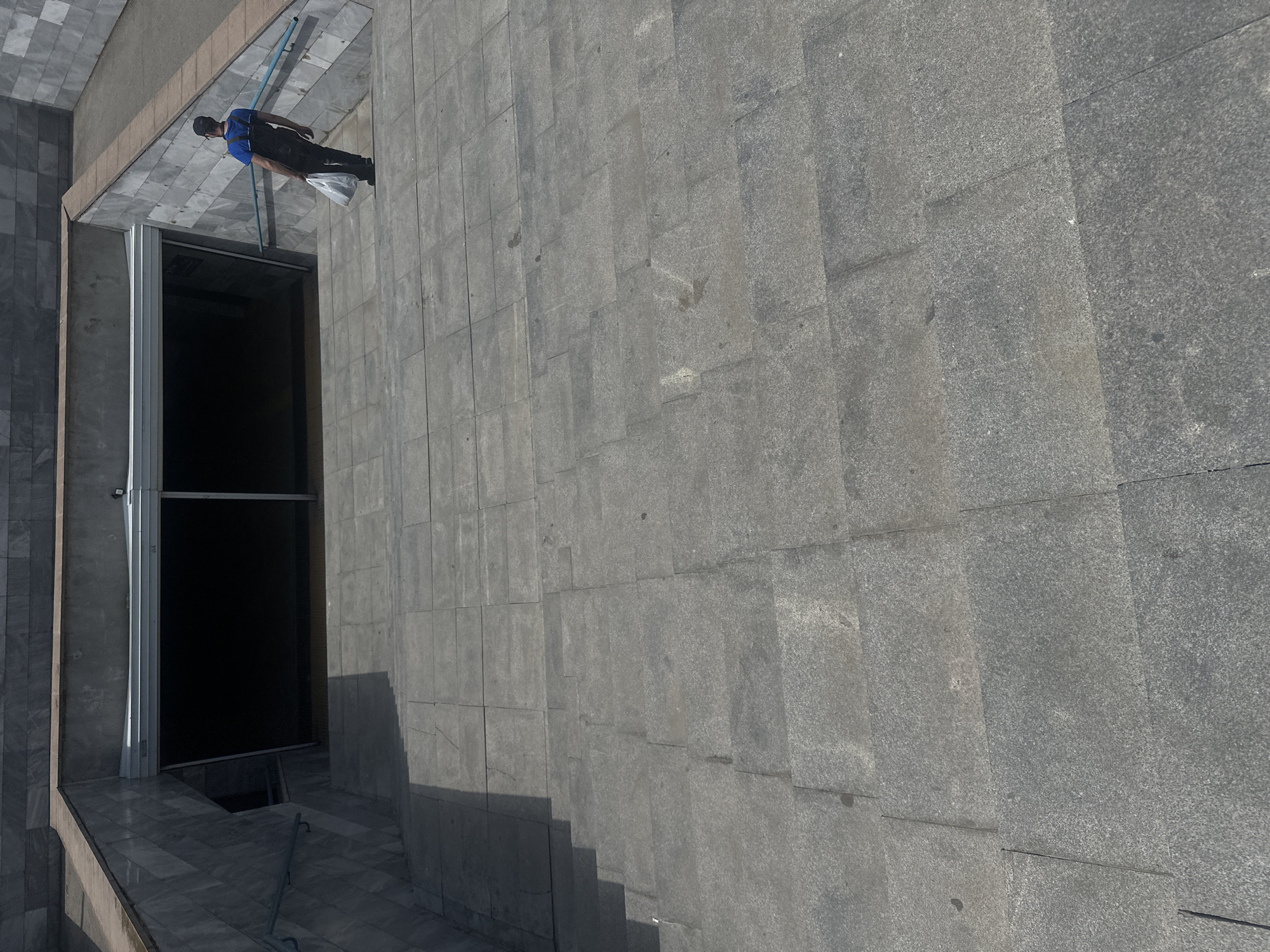 The image shows an outdoor stairway leading down into a dark underpass. The stairs and walls are constructed of gray stone tiles. A person wearing a blue shirt, dark overalls, and a cap is walking down the stairs, carrying a white plastic bag. The underpass entrance is covered by a metal roller shutter, partially open, revealing darkness within. A blue handrail runs along the side of the stairs. The scene appears to be in a public urban space, possibly a pedestrian underpass or subway entrance.