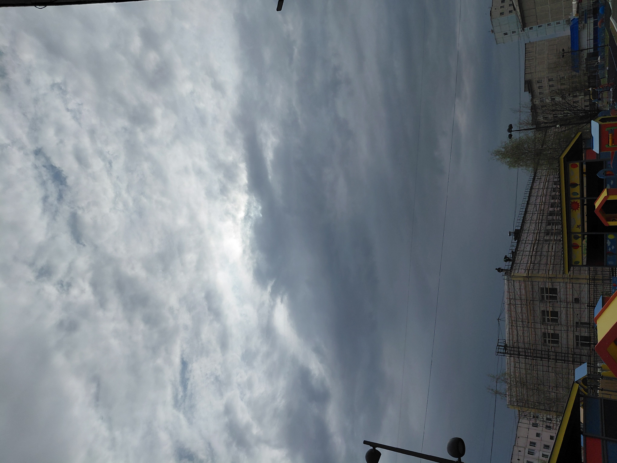 The image depicts an overcast sky over what appears to be a construction site adjacent to a playground. The location is Nadym, Russia, based on contextual clues such as architectural style. The sky is filled with gray and white clouds, with a hint of sunlight breaking through. A multi-story building is under construction and is covered in scaffolding. A playground is partially visible in the lower right corner, featuring colorful structures. A lamppost is in the foreground, its light bulb turned off. There is no sign of people or animals.