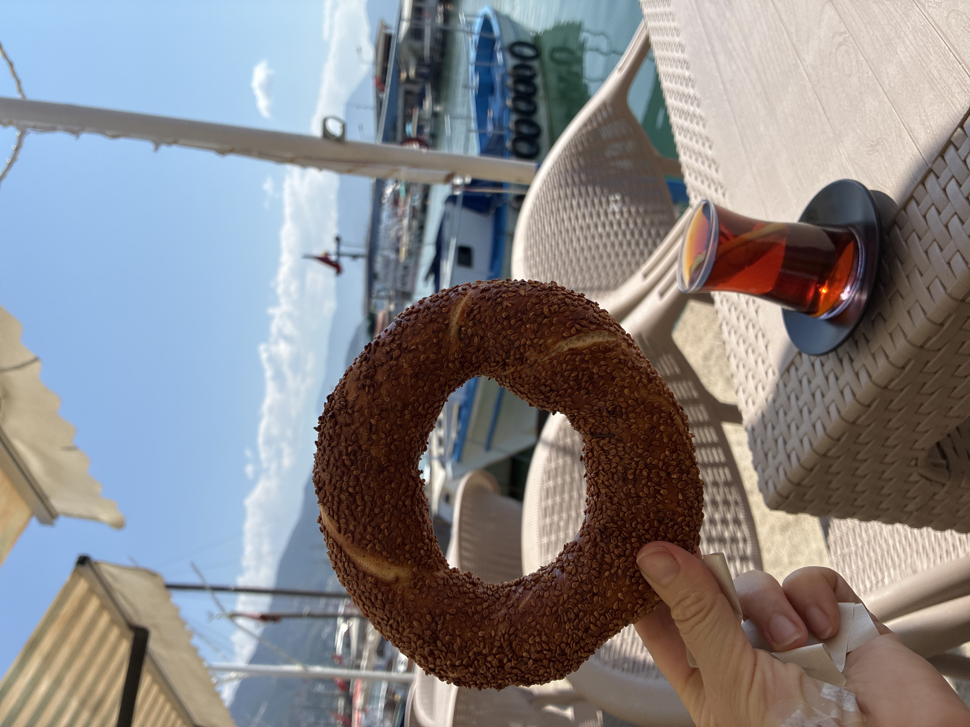An outdoor cafe scene in Alanya, Turkey, features a hand holding a simit (sesame-covered bread ring) in the foreground. To the right, a traditional tulip-shaped glass of Turkish tea sits on a saucer on a wicker table. The background reveals a harbor with several boats, calm blue water, and hazy mountains under a clear blue sky with scattered clouds. A striped awning is visible overhead. The ambiance suggests a relaxed, sunny day at a waterfront establishment.