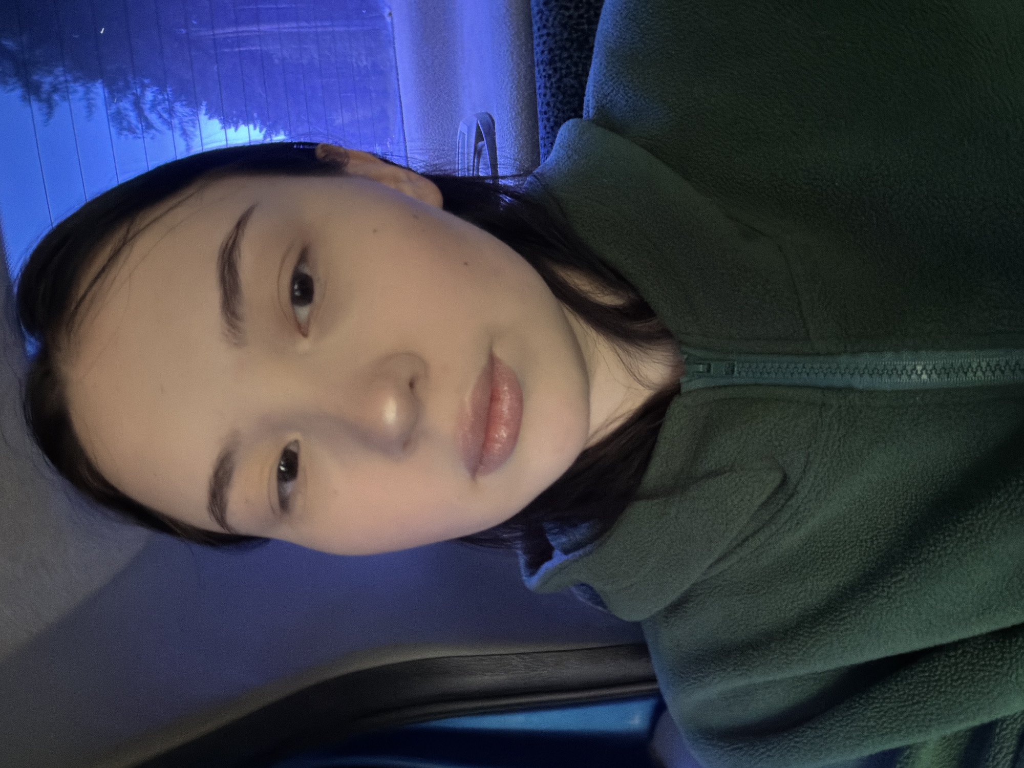 A close-up portrait shows a young person inside what appears to be a vehicle. The individual, with dark hair and a fair complexion, looks directly towards the camera with a calm and neutral expression. Their dark eyes are visible, along with well-defined eyebrows and softly colored lips. They are wearing a dark green or deep teal fleece jacket with a visible zipper on the front, extending from the neck.
The background, visible through what seems to be a car window, is dominated by a deep blue hue. This blue light suggests an evening or nighttime setting, with indistinct shapes and reflections, possibly from streetlights or illuminated buildings on a wet surface, creating vertical lines and patterns. Part of the car's interior, likely a headrest or seat, is also visible in muted tones in the lower and upper left of the frame. The overall lighting casts a soft glow on the person's face. No discernible text is visible in the image. The location context provided is Chaikovsky, Russia.