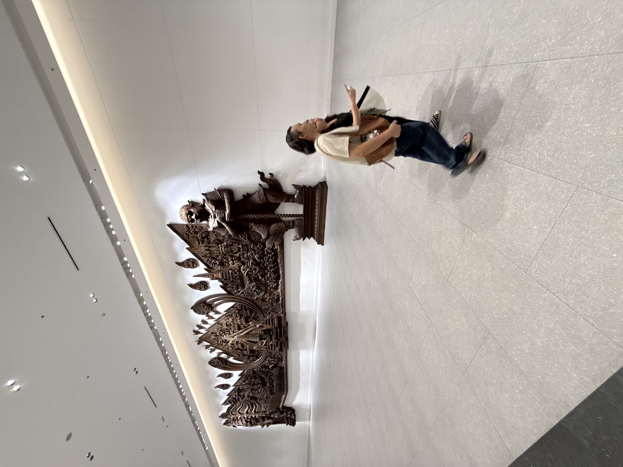 Two individuals, a man and a woman, are walking on the polished tile floor of a modern indoor space, possibly a museum or airport concourse, in Bangkok, Thailand. The man, wearing a light-colored t-shirt and dark pants, is in motion, his feet blurred. He carries a brown bag over his shoulder. The woman, in a dark top and dark pants, is walking beside him, gesturing with her right hand. Behind them, a large, intricate wooden relief sculpture adorns a white wall. The sculpture depicts mythological figures and architectural elements, with a prominent standing figure holding a sword in the center. The space is brightly lit by overhead lights, with a warm band of light illuminating the top of the wall behind the sculpture. The floor is tiled with large, light-colored tiles.