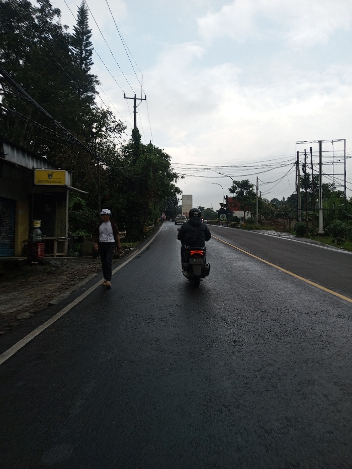The scene depicts an overcast day on a wet asphalt road in Dawathang_Dorjibi_Kashingtsawa, Bhutan. In the foreground, a dark-colored scooter, featuring a red taillight, is traveling away from the camera on the right side of the road. The operator wears dark clothing and a dark helmet with a white "007" logo on the back. To the left, a male pedestrian walks in the same direction, wearing a white t-shirt with brown accents, dark trousers, light-colored shoes, and a white baseball cap worn backward.
The left side of the road features a raised dirt/concrete shoulder alongside a single-story building with a blue roll-down shutter and a yellow sign above displaying the word "ROONUNCIA" and a cup-like symbol. A small wooden table or bench is visible outside this structure. Dense green foliage, including a tall coniferous tree, lines this side of the road. Overhead, multiple utility poles with a complex network of black electrical or communication wires traverse the sky, which is heavily clouded.
The road itself is dark and visibly wet, suggesting recent precipitation, and is delineated by a white solid line on the left and a faded yellow line on the right. In the distance on the right, a large, empty metal billboard frame stands prominently. Further down the road, additional white vehicles are discernible, along with a structure featuring a red roof and a dark monument or statue in front of it.