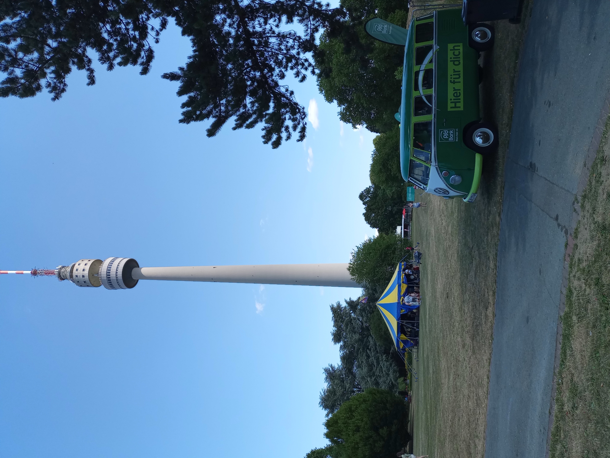 The image depicts the iconic Florian Tower in Dortmund, Germany, standing tall against a clear blue sky. In the foreground, a vibrant green vintage Volkswagen van, branded with the logo "PSD BANK Rhein-Ruhr eg" and the slogan "Hier für dich," is parked on a grassy area. To the left of the van, a yellow and blue striped tent is set up, with several people gathered around it. The ground is covered in dry, golden grass, suggesting a summer day. Trees are visible in the background, framing the scene, with the dense foliage of a pine tree on the right side of the image. The atmosphere appears to be that of an outdoor event or festival.