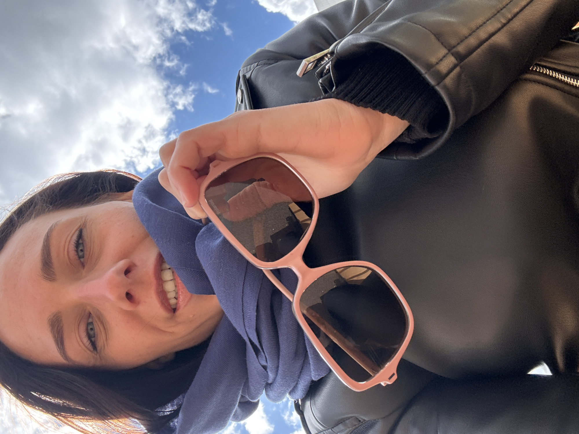 A woman with short dark hair and fair skin smiles towards the camera while holding pink-framed sunglasses. She wears a navy blue scarf and a black leather jacket. The sky with scattered clouds is visible in the background. The scene is likely Saint Petersburg, Russia, based on the style of the buildings reflected in the sunglasses.