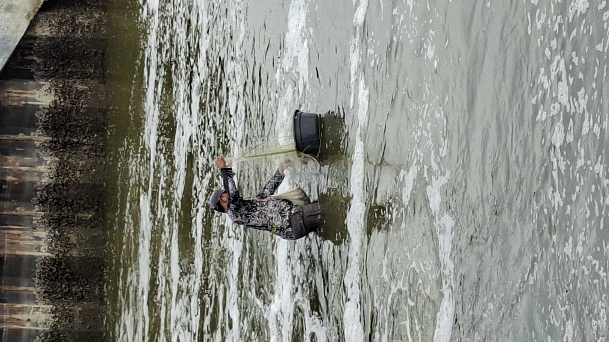 A person is fishing in the water near a docked structure. The person is wearing a cap, a patterned long-sleeved shirt, and dark pants. They are standing in the water, which is murky and has white foam on the surface. In their right hand, the person holds a fishing net, and they are using it to scoop something from the water into a dark plastic bucket placed beside them. A beige bag hangs from their left shoulder. The background shows the base of a structure, possibly a pier or a wall, with numerous small, dark objects attached to it, resembling mussels or barnacles. The water is calm with some ripples and currents. The scene appears to be a coastal or riverine environment. Based on the style of the docked structure and the type of fishing, this scene is likely in Lhokseumawe, Indonesia, where such activities are common along the coast and in river areas.