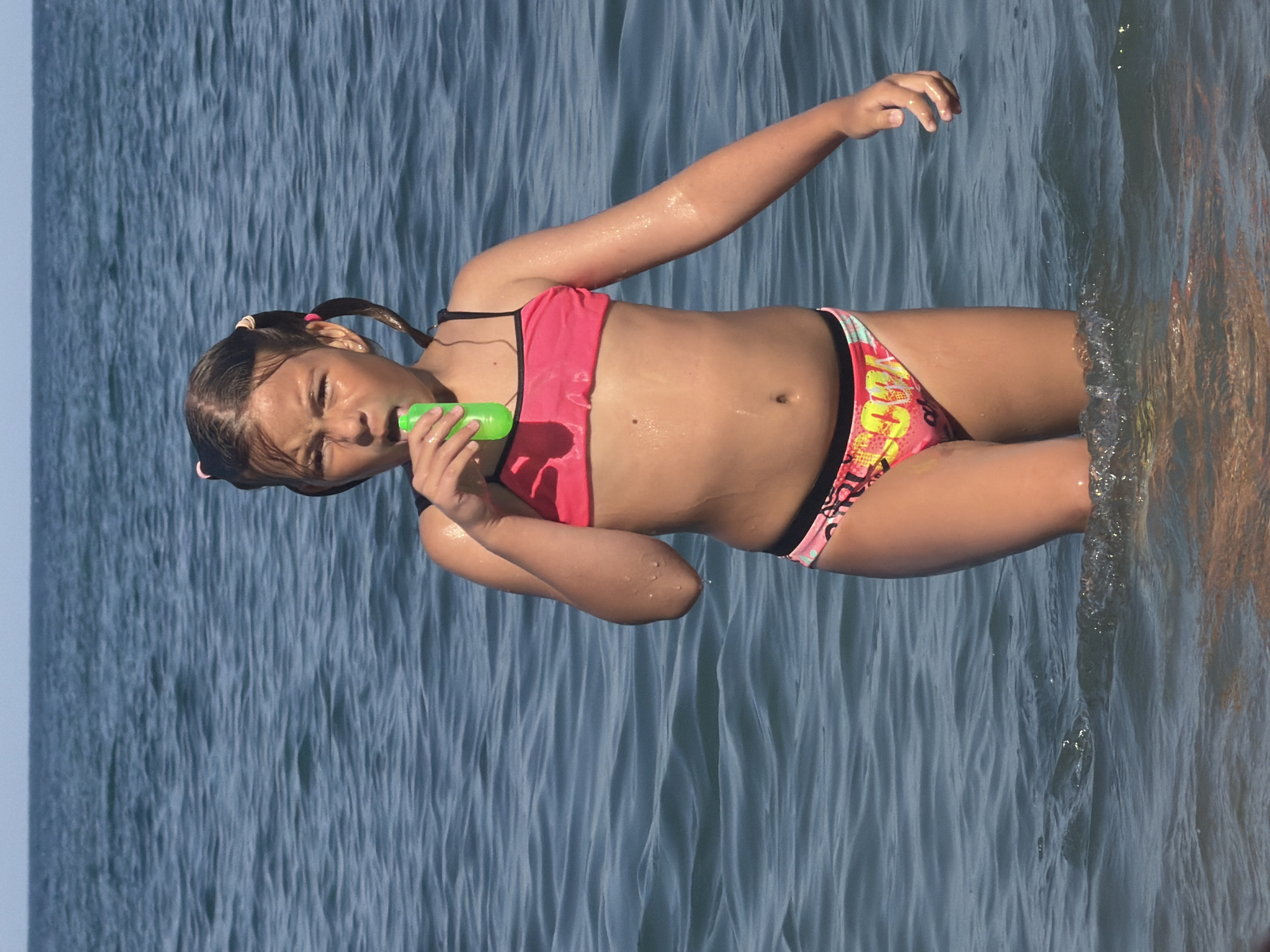 A young girl is standing in the sea, holding a green toy. She is wearing a pink bikini and has her hair in two pigtails. The water is blue and choppy, and the sun is shining. The girl looks to be having fun, with a playful expression on her face. The background is a blur of blue water, suggesting the vastness of the sea. The lighting indicates it is daytime, likely sunny. The image captures a moment of childhood joy and summer fun. The location is identified as Partenit, Ukraine. There is no visible text in the image.