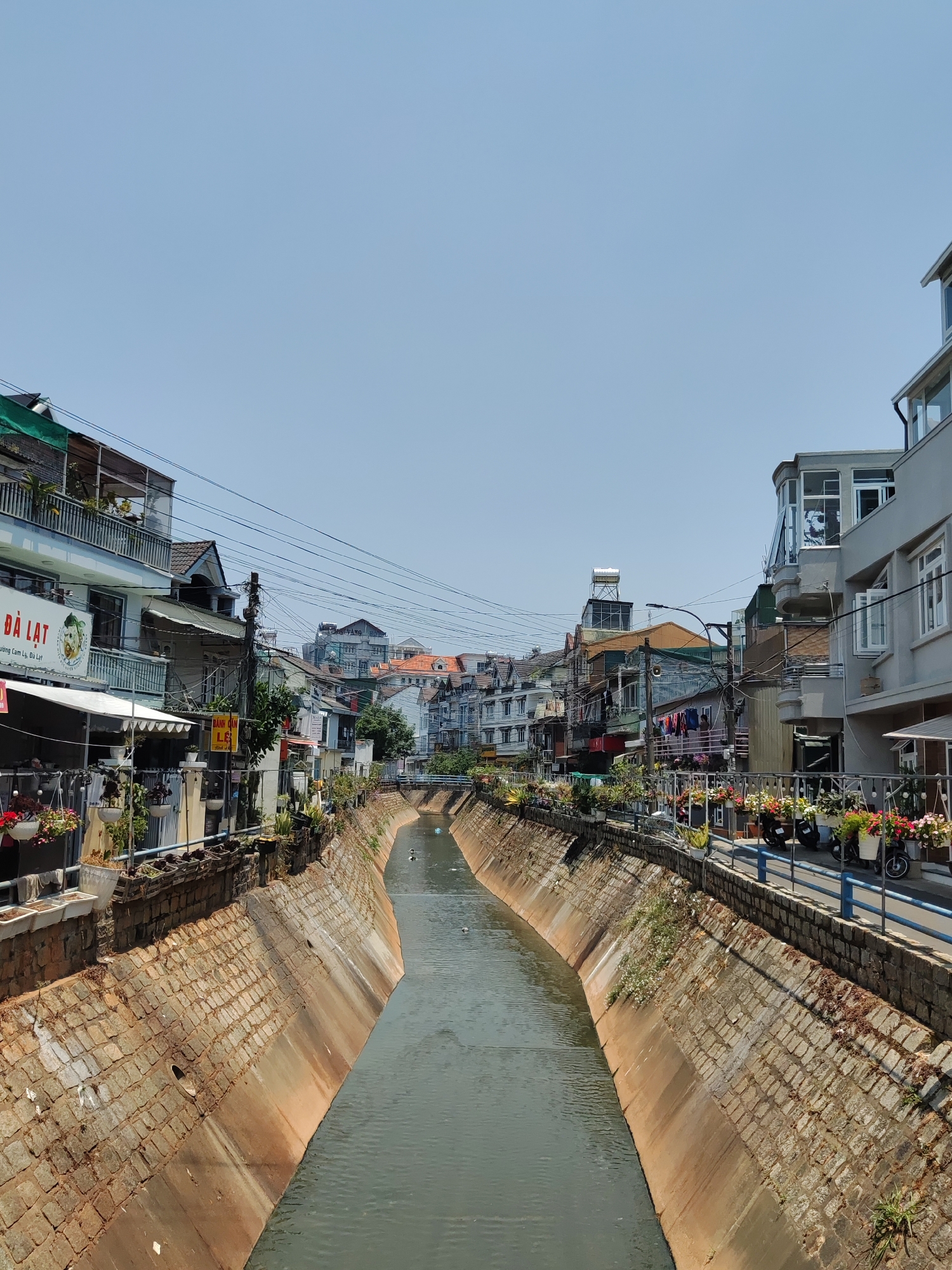 A canal runs through the city of Cam Ly, Đà Lạt, Vietnam, flanked by buildings on either side. The canal's banks are constructed of stone bricks that slant downwards to the water. Buildings of varying heights and architectural styles line the streets on both sides of the canal. Power lines crisscross the sky above the buildings. The sky is clear and blue. Flowers and plants are visible on the sidewalks and balconies along the canal. Several motorcycles are parked along the canal side.
