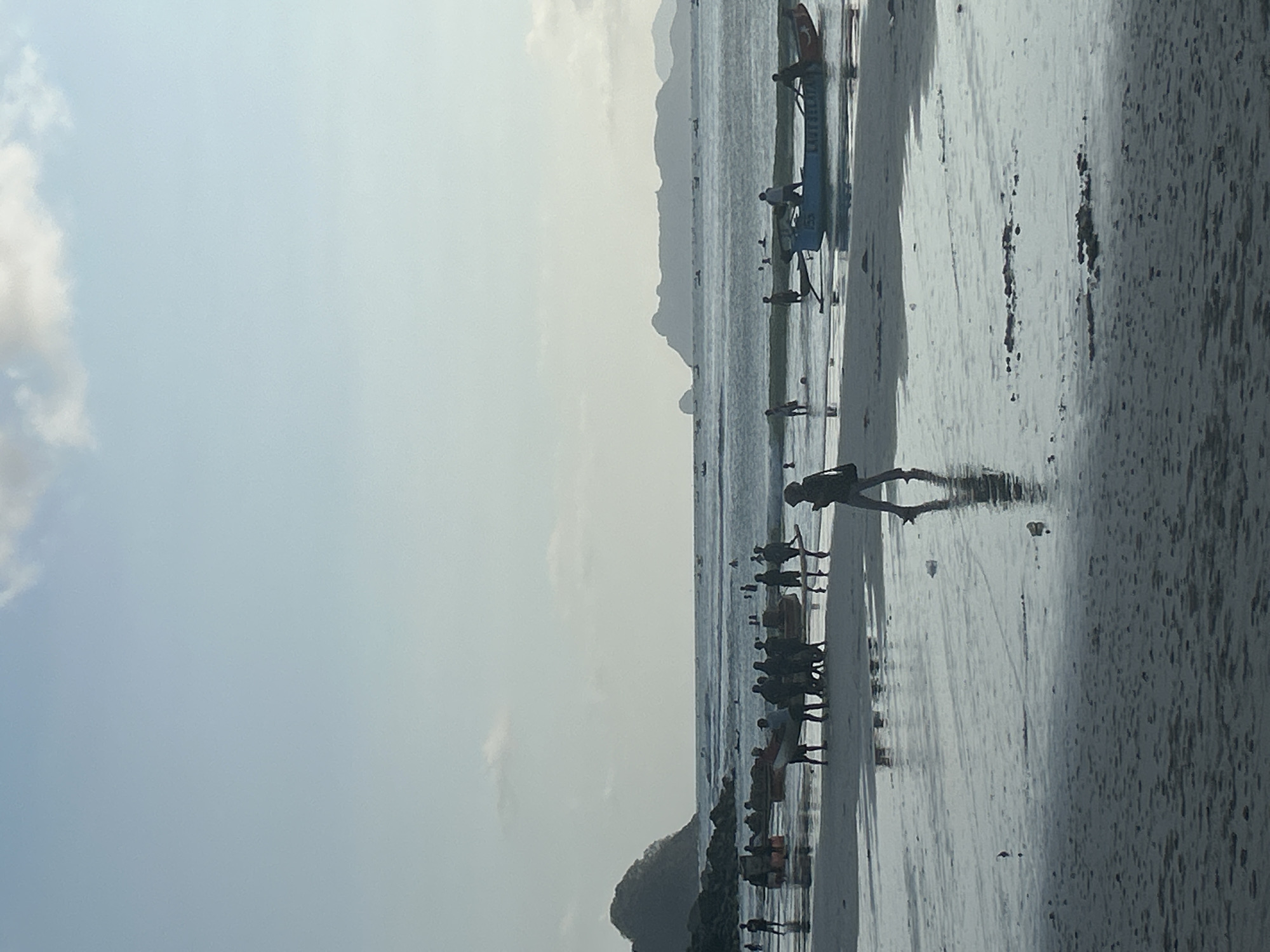 In Selongbelanak, Indonesia, a beach scene unfolds under a clear, pale blue sky with sparse clouds. The foreground is a wet, sandy beach with reflections of people and objects. Several groups of people are present on the beach and in the shallow water. In the middle ground, a group of people are gathered around several boats, some of which appear to be beached or being prepared. A lone figure walks across the wet sand in the mid-right of the frame, their reflection clearly visible. Further out, the ocean stretches towards a distant, hazy coastline with rolling hills and mountains. Several small boats are scattered across the water. The overall impression is a lively, coastal scene with people engaged in various beach and boating activities.