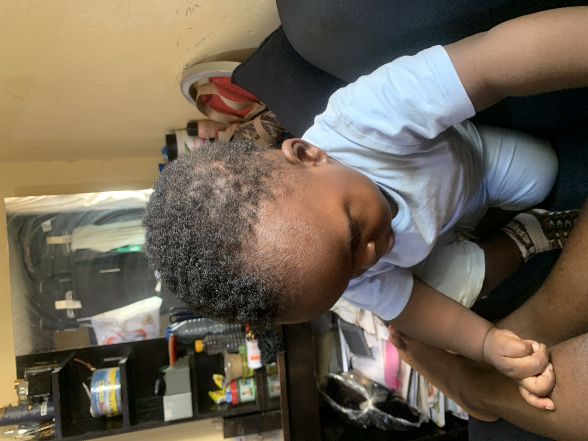 An overhead, medium shot shows a young Black child with curly hair, wearing a light blue t-shirt and light blue shorts, kneeling on a dark blue couch. The child is looking down and holding an adult's hand with their right hand. The adult's legs are visible in the foreground, and they appear to be dark-skinned. In the background, a shelf unit with various items is reflected in a mirror, and a yellow wall is visible. There are also some pillows on the couch. The scene is indoors and appears to be a living room.