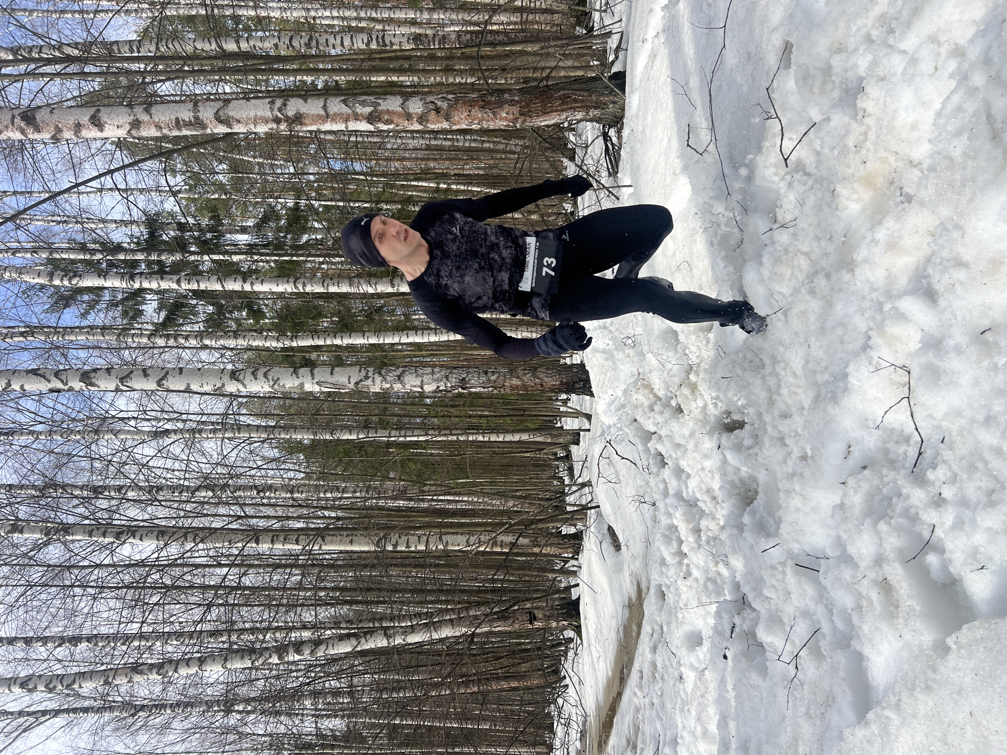 A man is positioned in a winter environment, possibly during a race or training session, based on his attire and race number "73." He is wearing black athletic clothing consisting of a beanie, long-sleeved shirt, pants, and gloves. The man is standing on a snowy terrain. Behind him is a dense forest of birch trees, with their characteristic white bark and thin, bare branches reaching towards the sky. The sky is partially visible through the branches, appearing to be a light blue color. The presence of birch trees suggests this is in Russia, but there is no definitive evidence of the city of Zelenodolsk, Russia in the image.