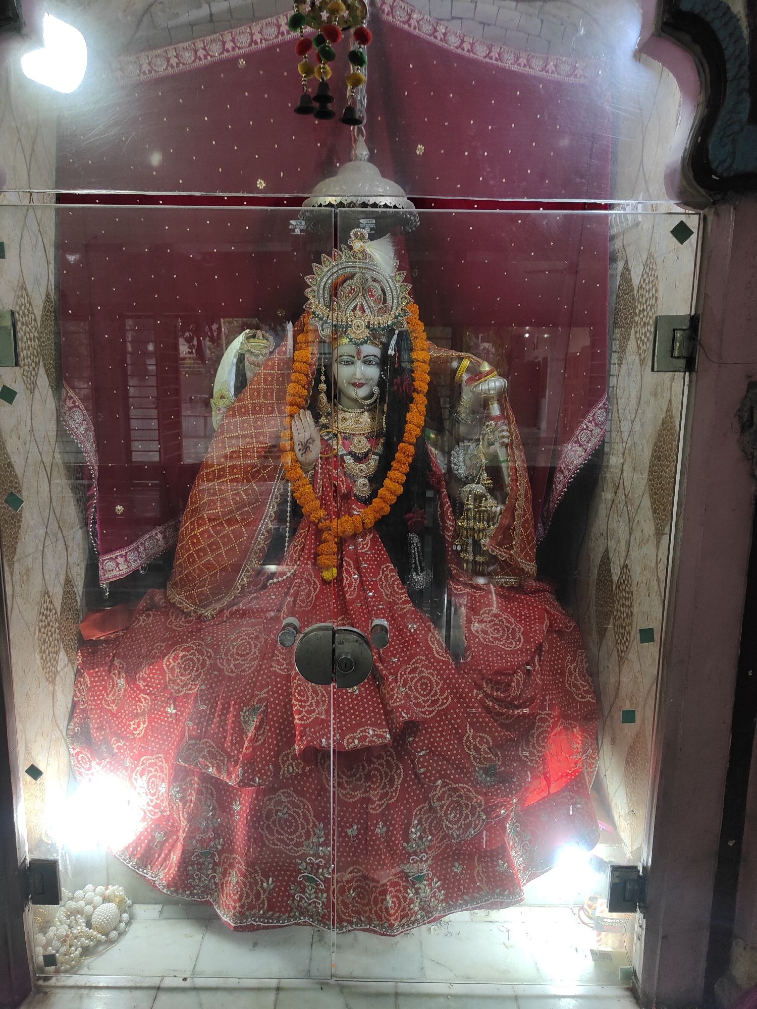 The image shows a statue of a Hindu goddess, likely Durga, housed in a glass-enclosed shrine. The statue is adorned with elaborate red and gold attire, orange marigold garlands, and intricate jewelry. The shrine is decorated with red fabric, and the background features ornate wall coverings. The lighting within the shrine is bright, with visible light bulbs. Outside the shrine, on the floor, are several strings of white beaded decorations. The overall scene suggests a religious setting, possibly a temple or a personal shrine. This shrine is located in Gurgaon, India.