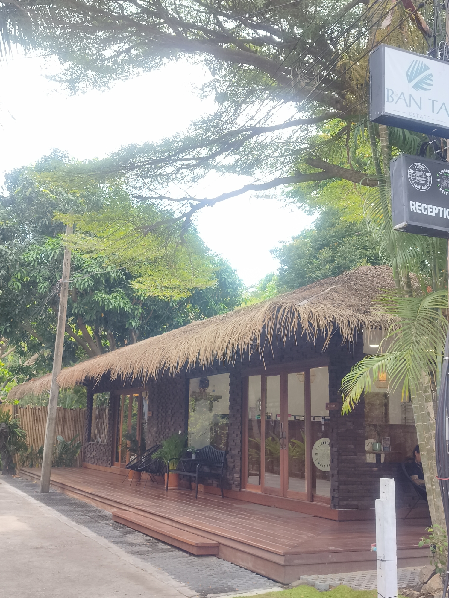 The image captures a building in Baan Tai, Thailand, with a thatched roof and wooden exterior. A sign above reads "BAN TAI ESTATE" and "RECEPTION". The building has a wooden deck with seating. Greenery surrounds the building, with trees visible in the background. A utility pole stands to the left of the building, and power lines run overhead. A palm tree is located to the right of the building. A person is visible sitting inside the building. The overall scene suggests a tropical or resort-like setting.