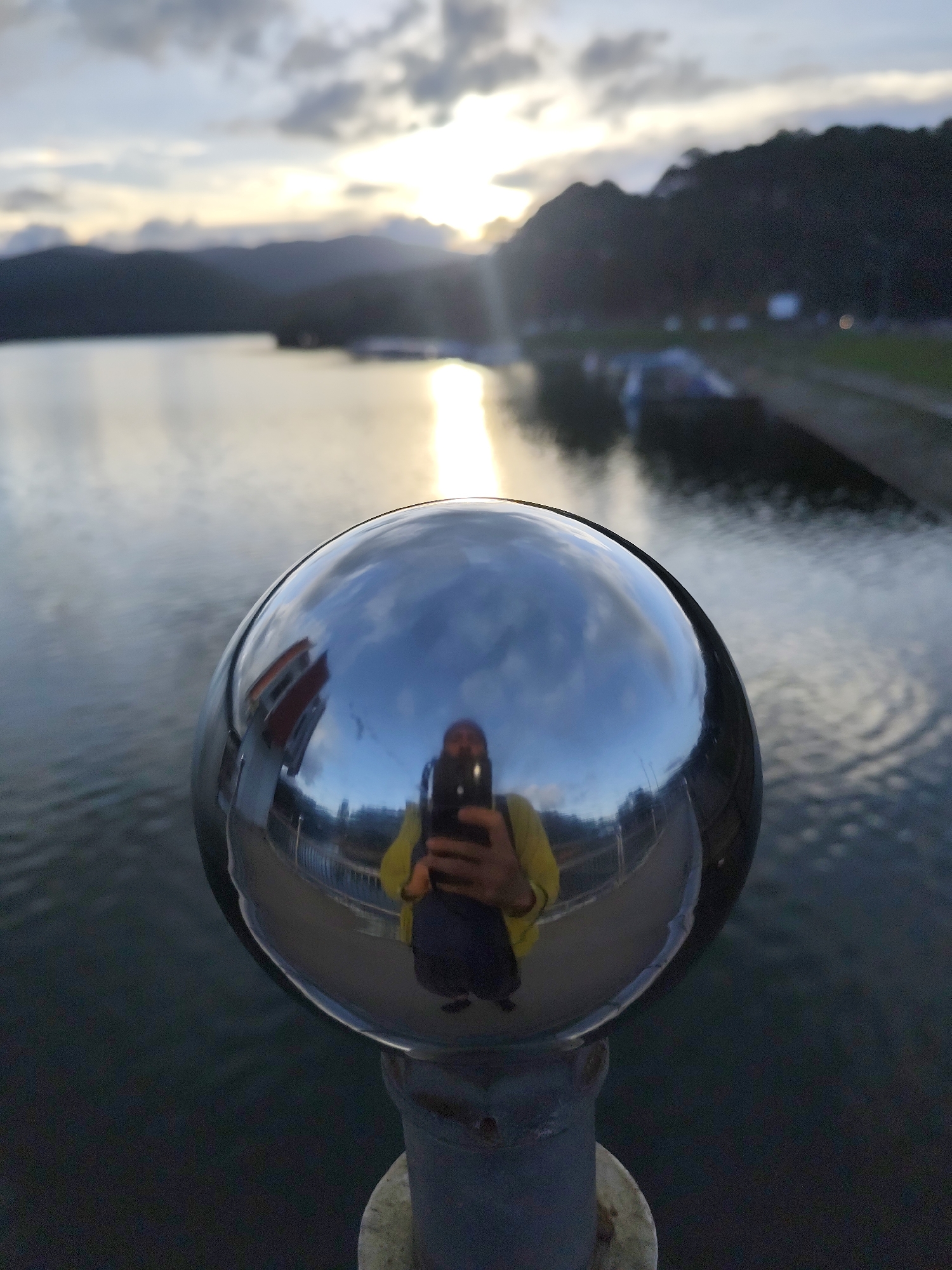 A medium shot captures a reflective spherical object perched atop a weathered, cylindrical post, with a body of water and distant mountains forming the backdrop. The reflection in the sphere shows the photographer, holding a smartphone, wearing a yellow shirt and dark pants. The sky is overcast with hints of sunlight breaking through the clouds, casting a reflection on the water. Along the water's edge, there is a bank with some indistinct objects, possibly boats or structures.