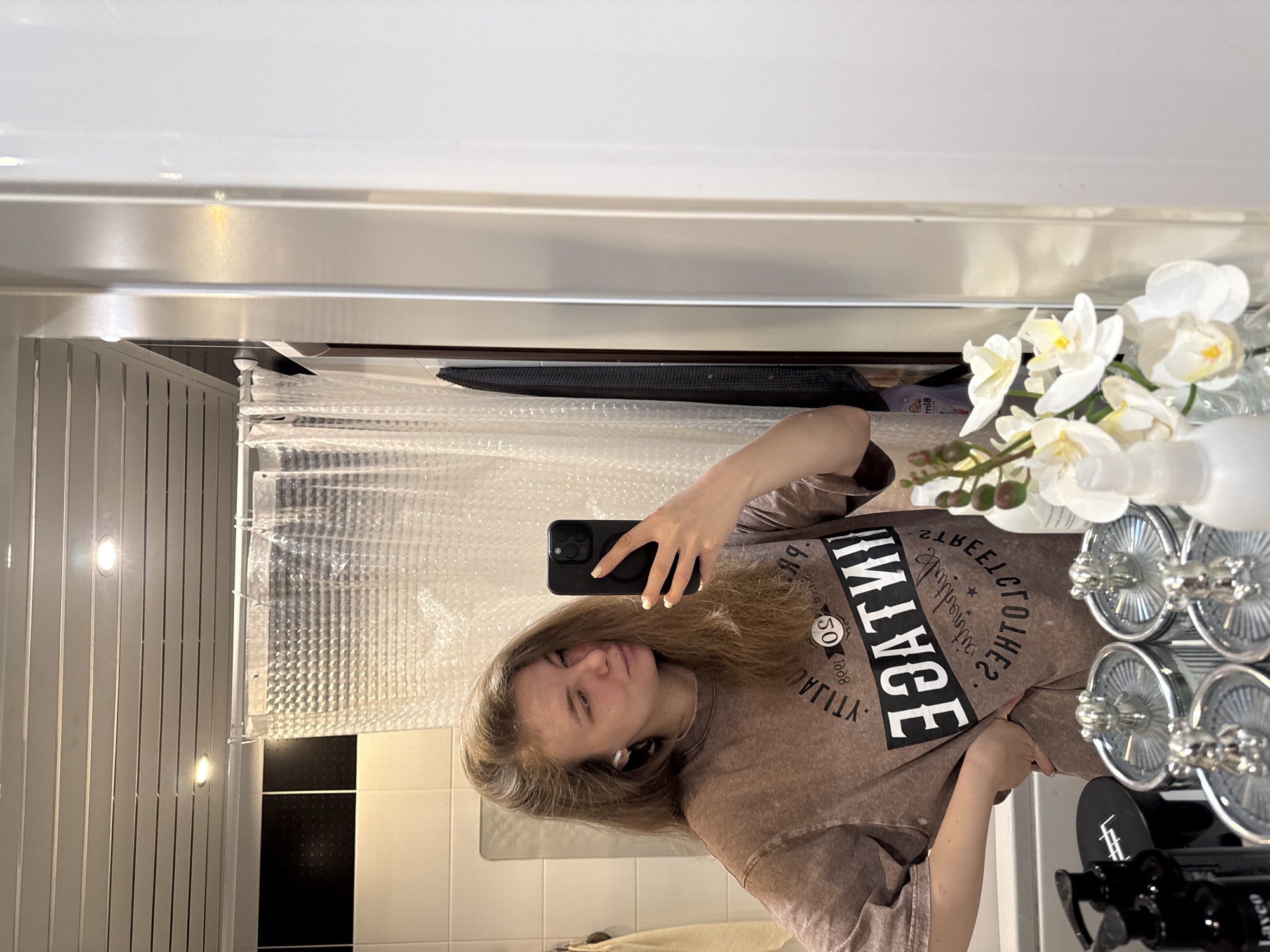 A woman is taking a selfie in a bathroom. The woman has blonde hair and is wearing a brown t-shirt. She is holding a black smartphone in her right hand. The bathroom has a shower with a clear plastic shower curtain, white tiles, and a black stripe. There are artificial flowers and cosmetic items on the counter. The location is possibly Vladimir, Russia.