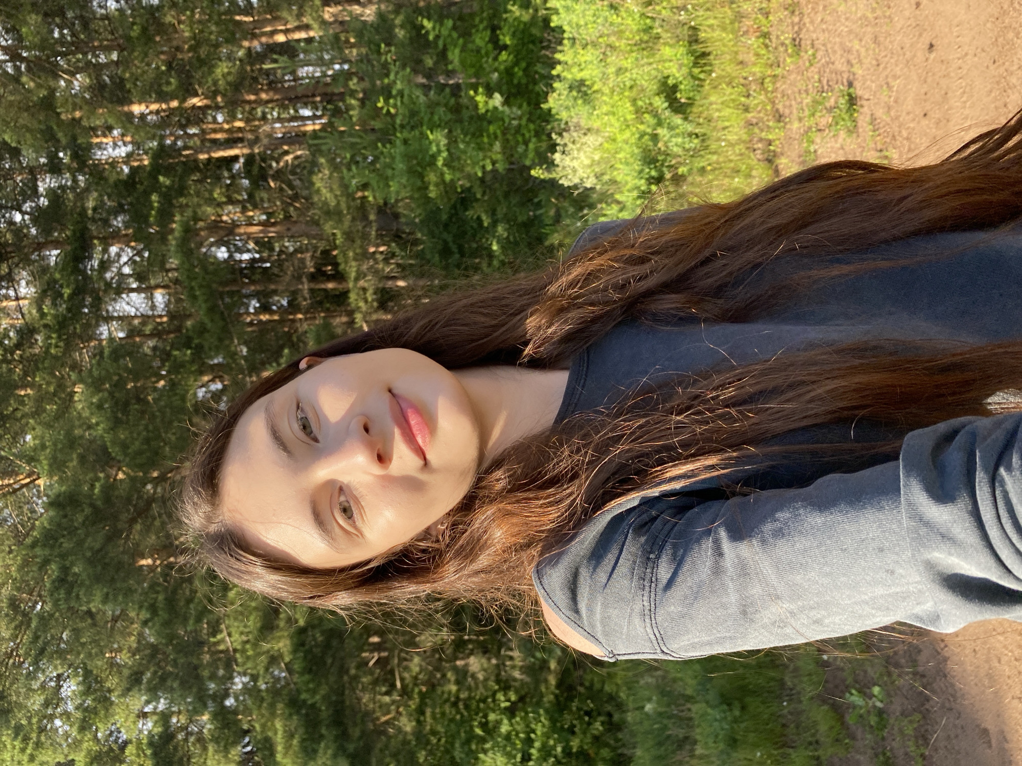A young woman with long, dark brown hair is shown in a selfie. She is outdoors, with a forest and a dirt path in the background. The sun is shining, casting a warm glow on her face and hair, suggesting it is either morning or late afternoon. The foliage in the background is lush and green, indicating a warm season. She is wearing a dark gray long-sleeved shirt. Her expression is relaxed and natural, with a slight smile. The image appears to be taken in Votkinsk, Russia. There is no visible text in the image.