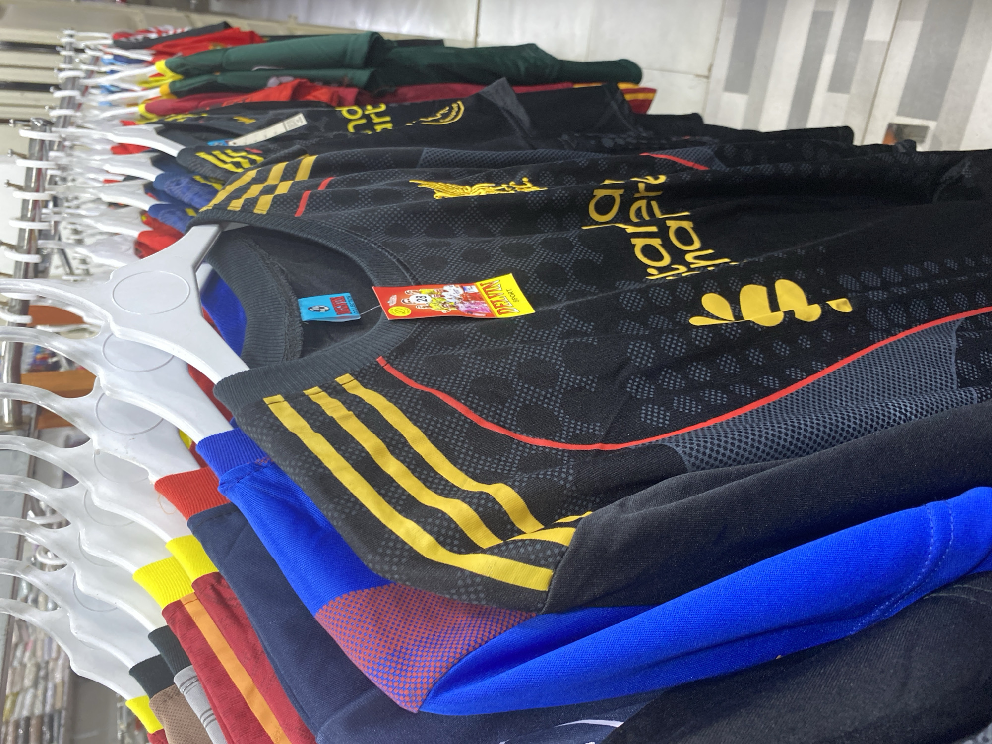 The image shows a row of sports jerseys hanging on white hangers on a metal rack. The jerseys are varied in color, with prominent black, red, yellow, and blue designs. Several jerseys feature yellow stripes and some have red accents. One black jersey in the foreground has a yellow crest and "LFC" lettering on it. Attached to some of the jerseys are colorful tags, one of which displays the brand name "DELVIN SPORT". The background appears to be an indoor retail environment with white shelving units visible. The lighting suggests an indoor setting, possibly a clothing store. The image is taken from a slightly elevated angle, looking down at the hanging garments. There are no people visible in the image. This scene is located in Lamteumen Barat, Indonesia.