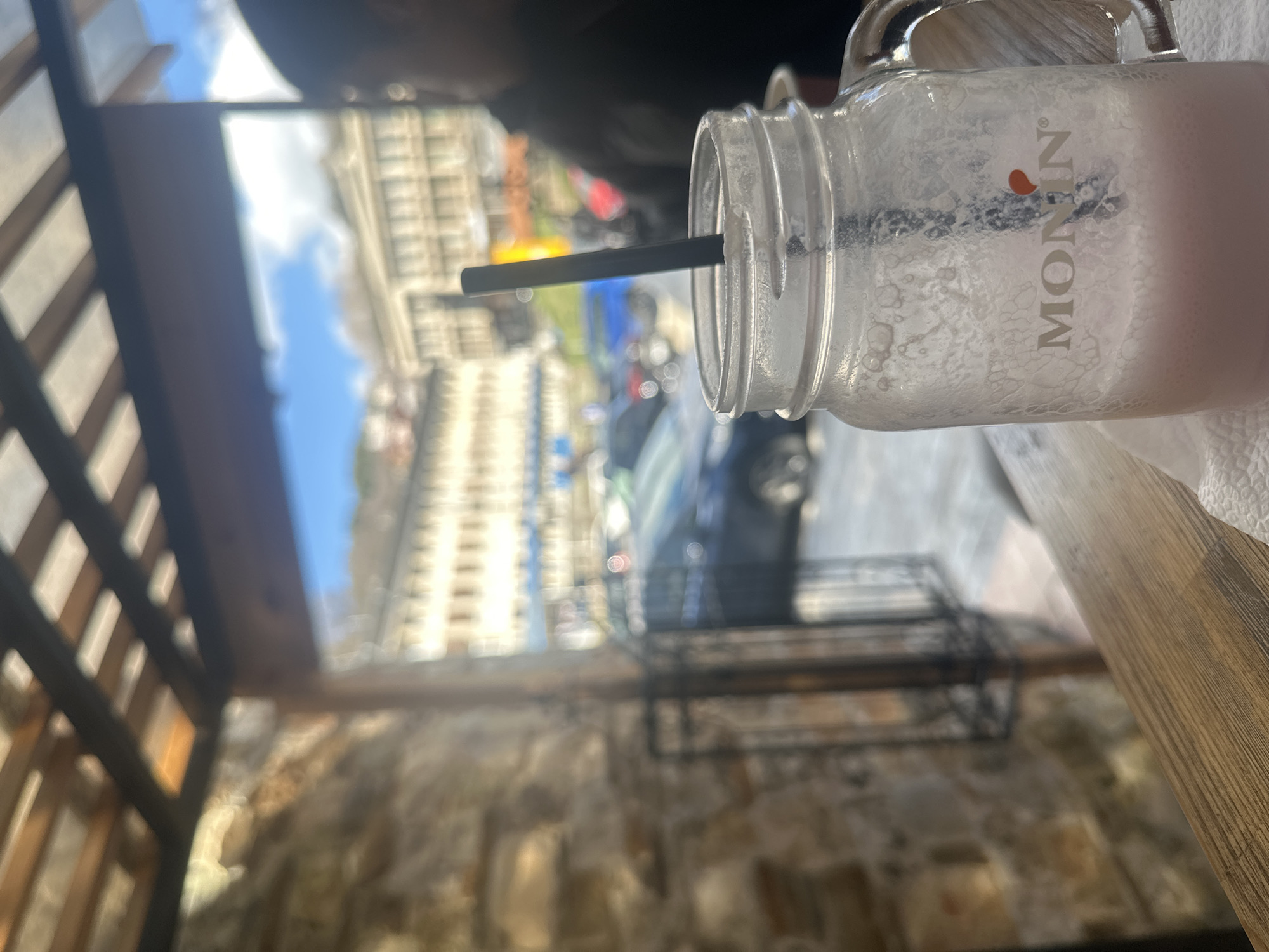 The photograph depicts a glass jar containing a light pink-colored beverage, likely a milkshake or smoothie, sitting on a wooden table. A black straw is inserted into the drink. The jar features the logo of "MONIN" printed on the side.
Through an out-of-focus window, a view of a city street is visible. Buildings line the street, and several vehicles are parked or moving along the road. The urban environment appears to be a blend of residential and commercial structures. Based on this image, the location is determined to be in Smolyan, Bulgaria. A person is present, but their face is out of focus. The photograph appears to have been taken from an elevated position, potentially a balcony or a restaurant with an outdoor seating area. There is a wooden trellis-like structure above and to the left. There is stonework in the lower left background.