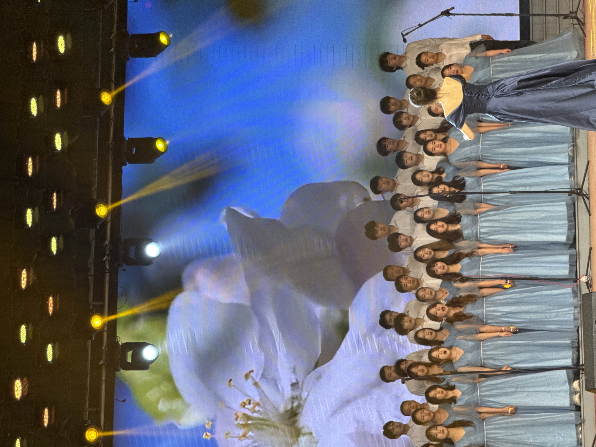 This media file captures a vibrant choral performance on an indoor stage, likely in Guta, China. The image is composed of a large choir, predominantly young adults and teenagers, arranged in several rows on risers, facing towards the front. Male choir members are dressed in white long-sleeved shirts, while female members wear light blue long dresses, some with a delicate shimmer.
A solo female performer, viewed from behind, stands prominently in the center-left. She is adorned in an elegant, dark blue strapless gown featuring a corset-style back, and her hair is styled in an updo with a decorative element. Microphones on stands are visible throughout the choir, indicating an active vocal performance.
The stage is illuminated by multiple bright stage lights positioned on the left side, casting beams of yellow and white light across the scene. The entire backdrop is a large projection screen displaying a beautiful, soft-focus image of a white flower, possibly an apple blossom, against a radiant blue background. Performers appear focused and engaged in the event, which suggests a formal concert, competition, or celebratory occasion. There is no visible text in the image.