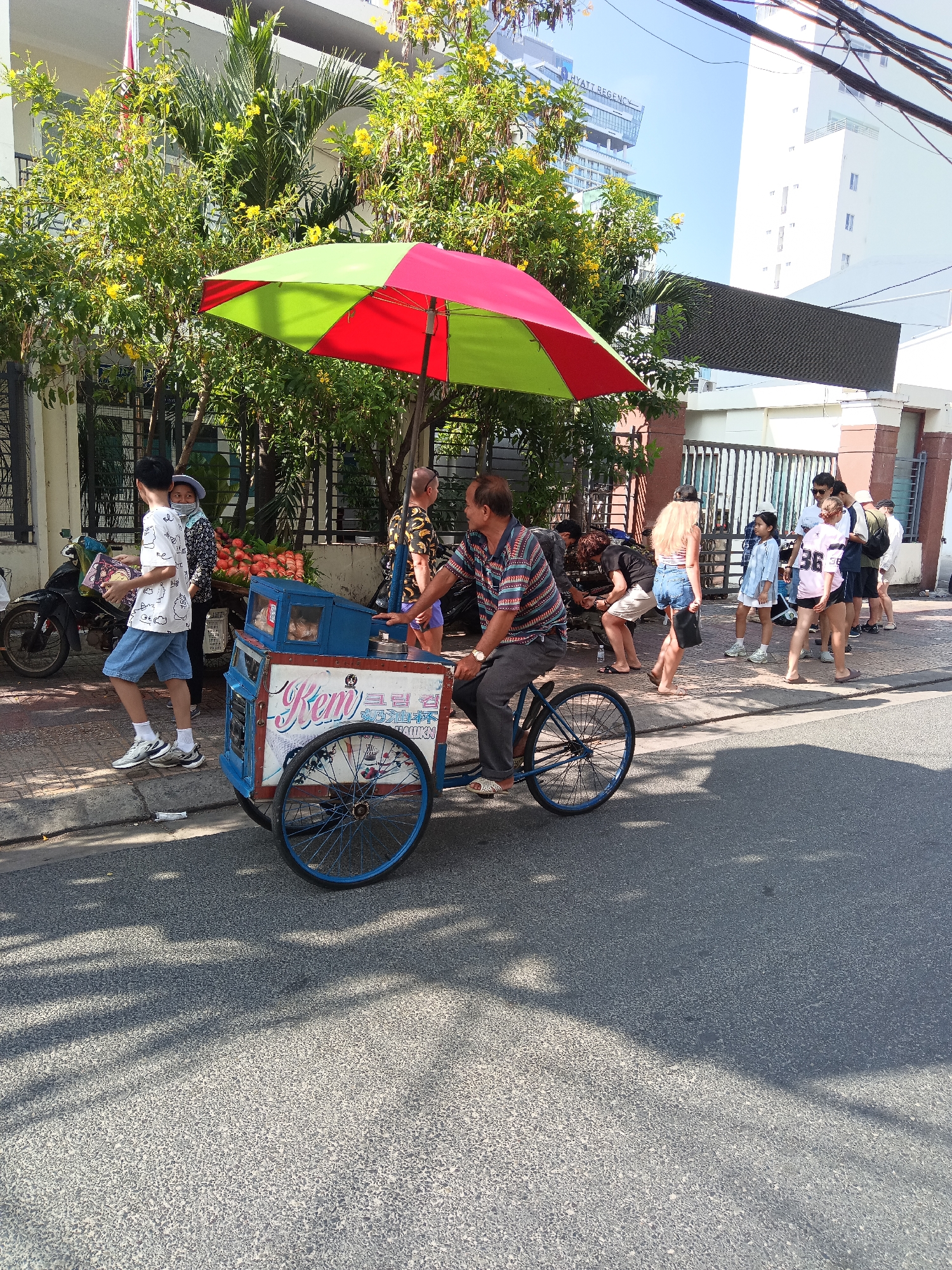 This is a street scene in Nha Trang, Vietnam, featuring a man pushing an ice cream cart. The cart is blue and white with text in what appears to be Vietnamese and Korean script. A red and green umbrella shades the cart. Several people are walking along the sidewalk, including a young boy in a white t-shirt and denim shorts, and a woman wearing a headscarf and carrying produce. In the background, there are buildings and trees. The street is paved with asphalt and has a faded white line marking the edge. Shadows from trees and overhead wires are cast on the road.