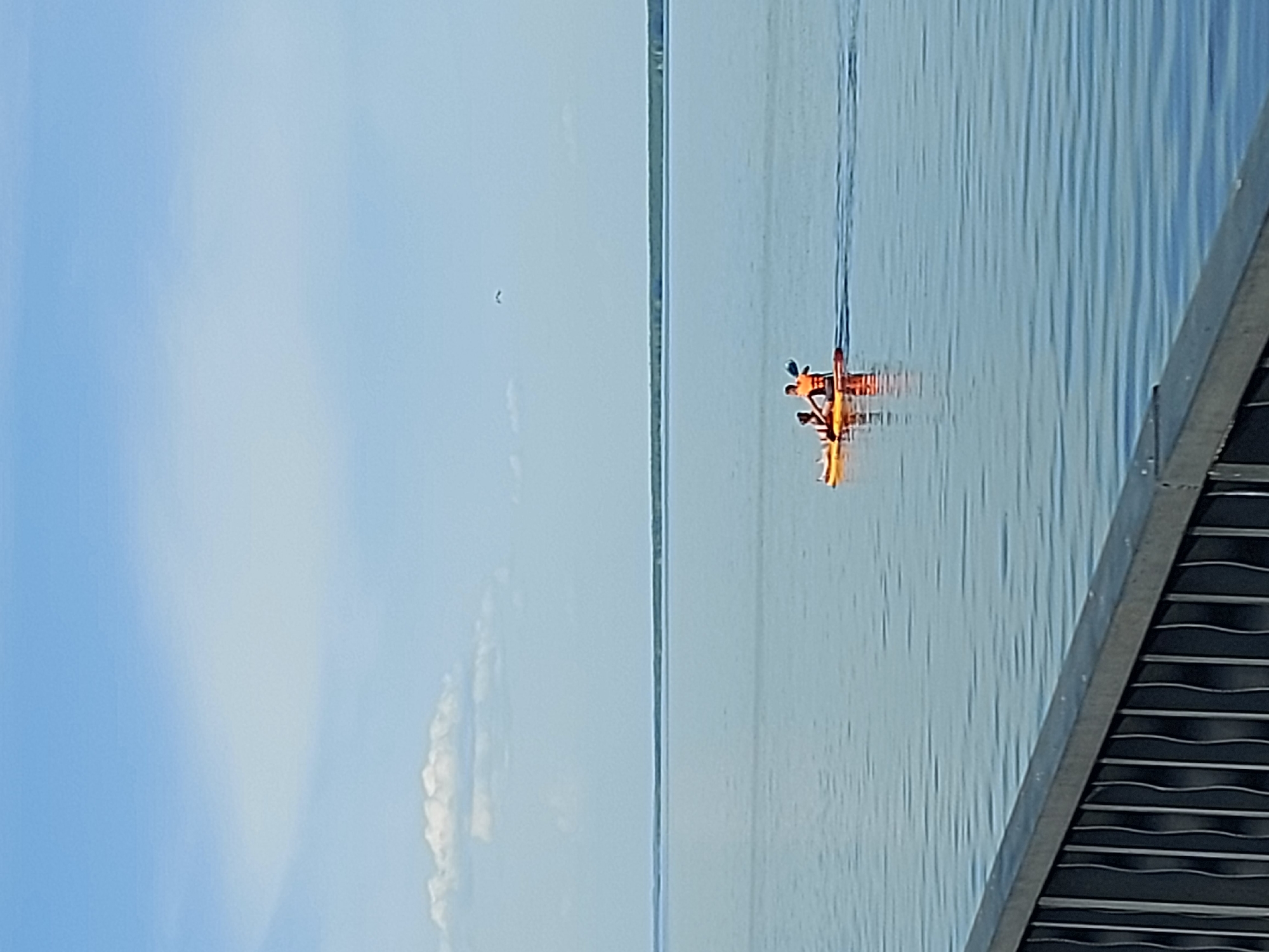The image depicts a body of water under a clear sky, possibly in the city of Chaikovsky, Russia. A yellow kayak with two people wearing orange life vests is visible on the water. Part of a metal structure with parallel lines is in the lower right corner.