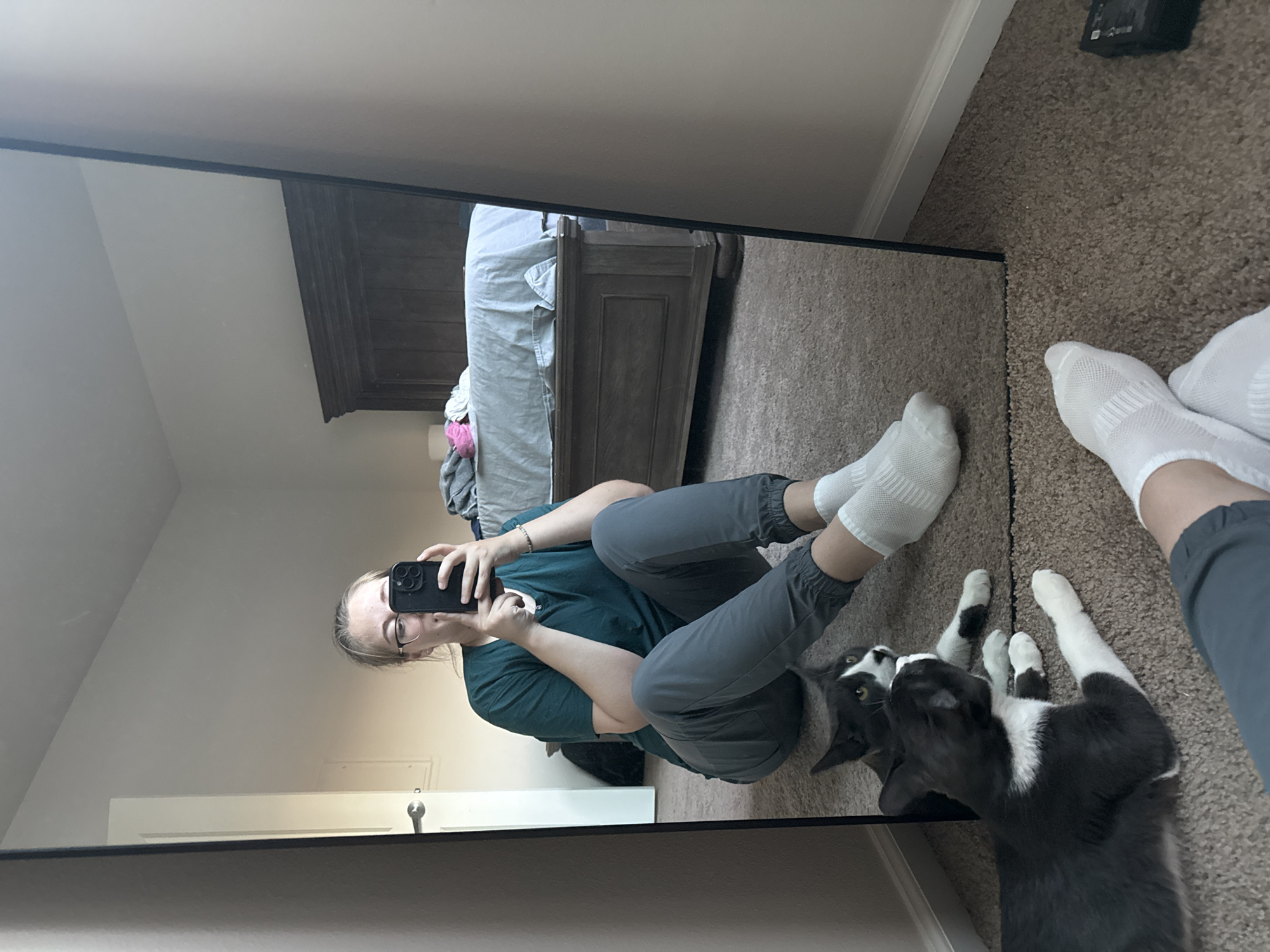 A person wearing a teal scrub top, grey jogger pants, and white socks is sitting on the floor in front of a full-length mirror, taking a selfie with a black smartphone. The person is wearing glasses and has blonde hair pulled back. Two cats, one grey and white, and another black and white, are lying on the floor near the person. The mirror reflects the person, the cats, a bed with a wooden headboard, and a door with a silver handle. The room has beige walls and a brown carpet. The image appears to be taken indoors in a residential setting, possibly in a bedroom. The metadata indicates the location as Fife, United States of America.